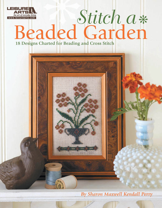 A framed cross-stitch with colorful seed beads forms a floral display beside a ceramic bird, blue thread spool, and vase with orange blooms. Above sits “Stitch a Beaded Garden—18 Designs Charted for Beading and Cross Stitch” by Leisure Arts.