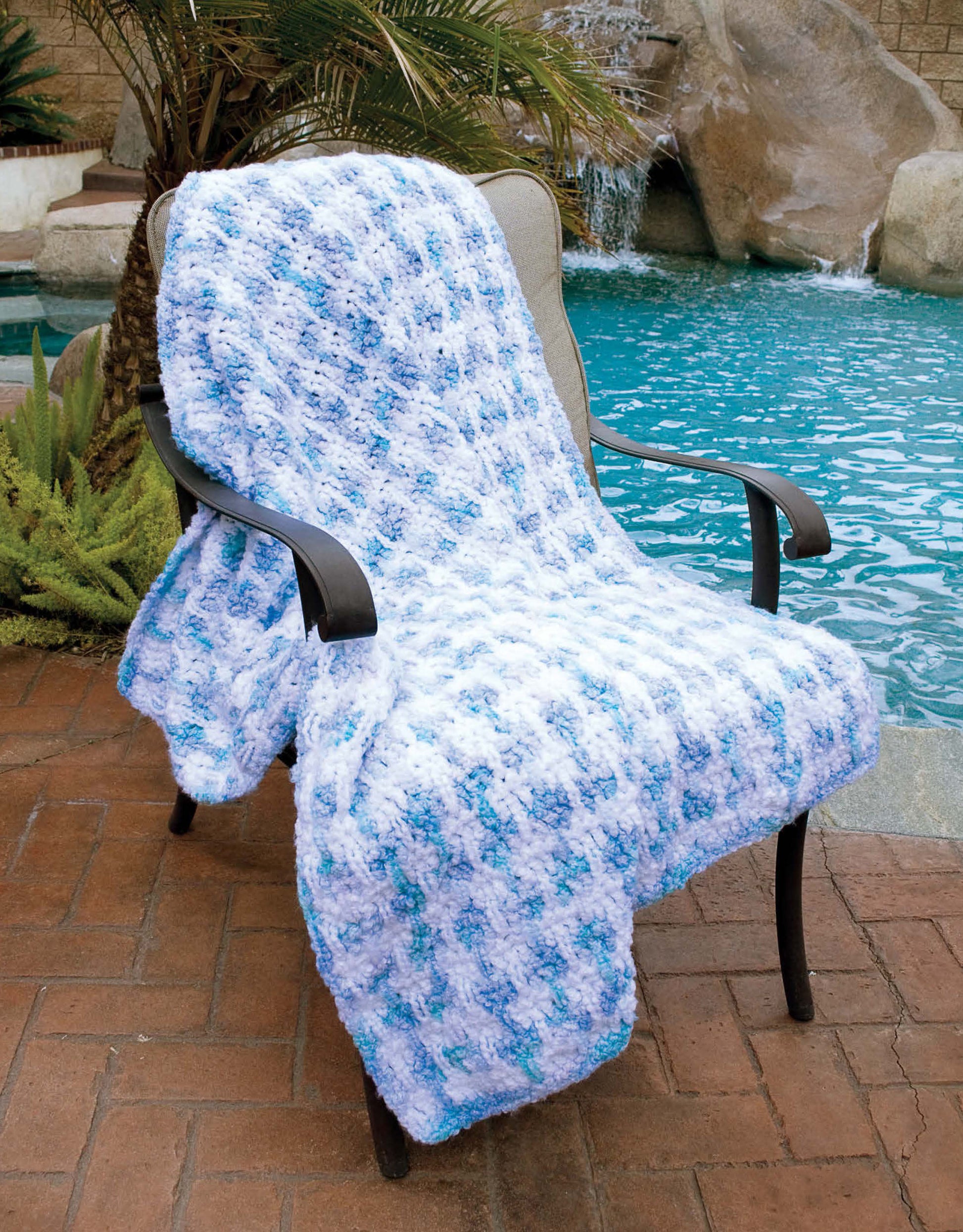 A cozy blue and white blanket from Leisure Arts’ More 48-Hour Afghans Digital Download drapes over a poolside chair, set against tropical plants and a waterfall backdrop.