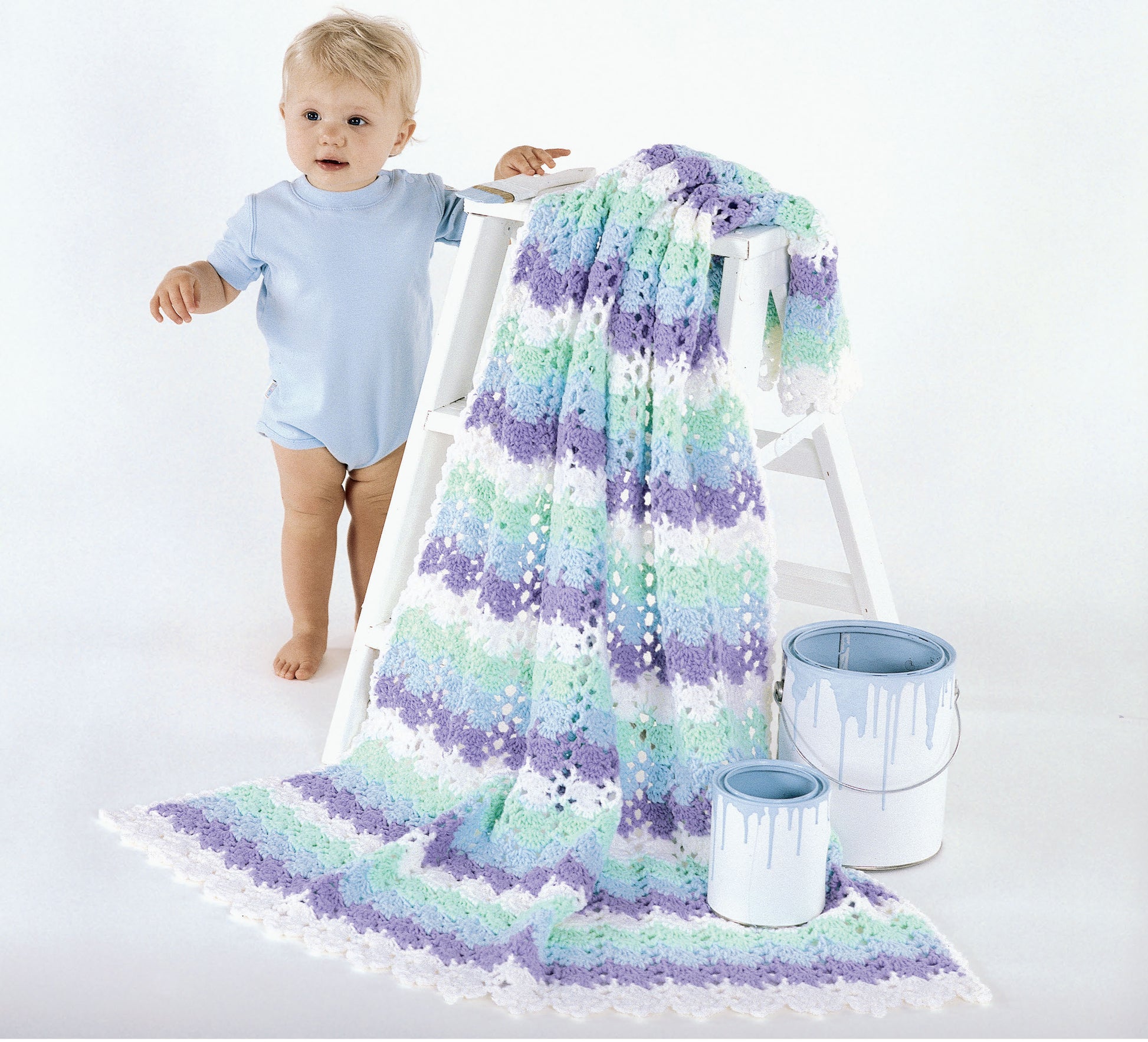 A baby in a light blue onesie stands by a white ladder draped with a pastel-striped crocheted blanket inspired by "The Big Book of Baby Afghans" from Leisure Arts. Two blue paint cans sit nearby against the white background.