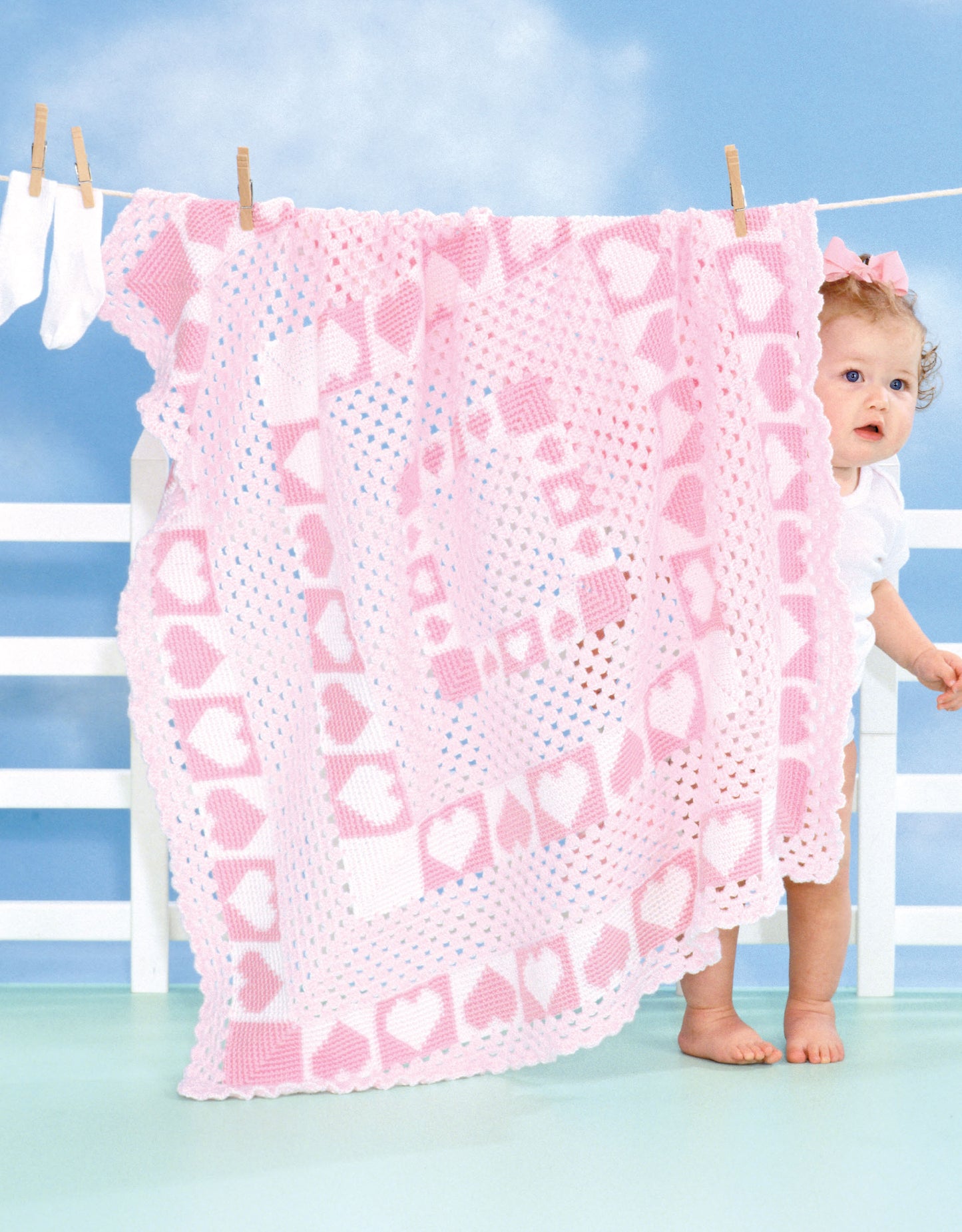 A baby in white stands by a clothesline, peeking from behind a pink and white crocheted afghan with hearts—one of the charming patterns featured in "The Big Book of Baby Afghans" by Leisure Arts—drying against a bright sky and white fence.