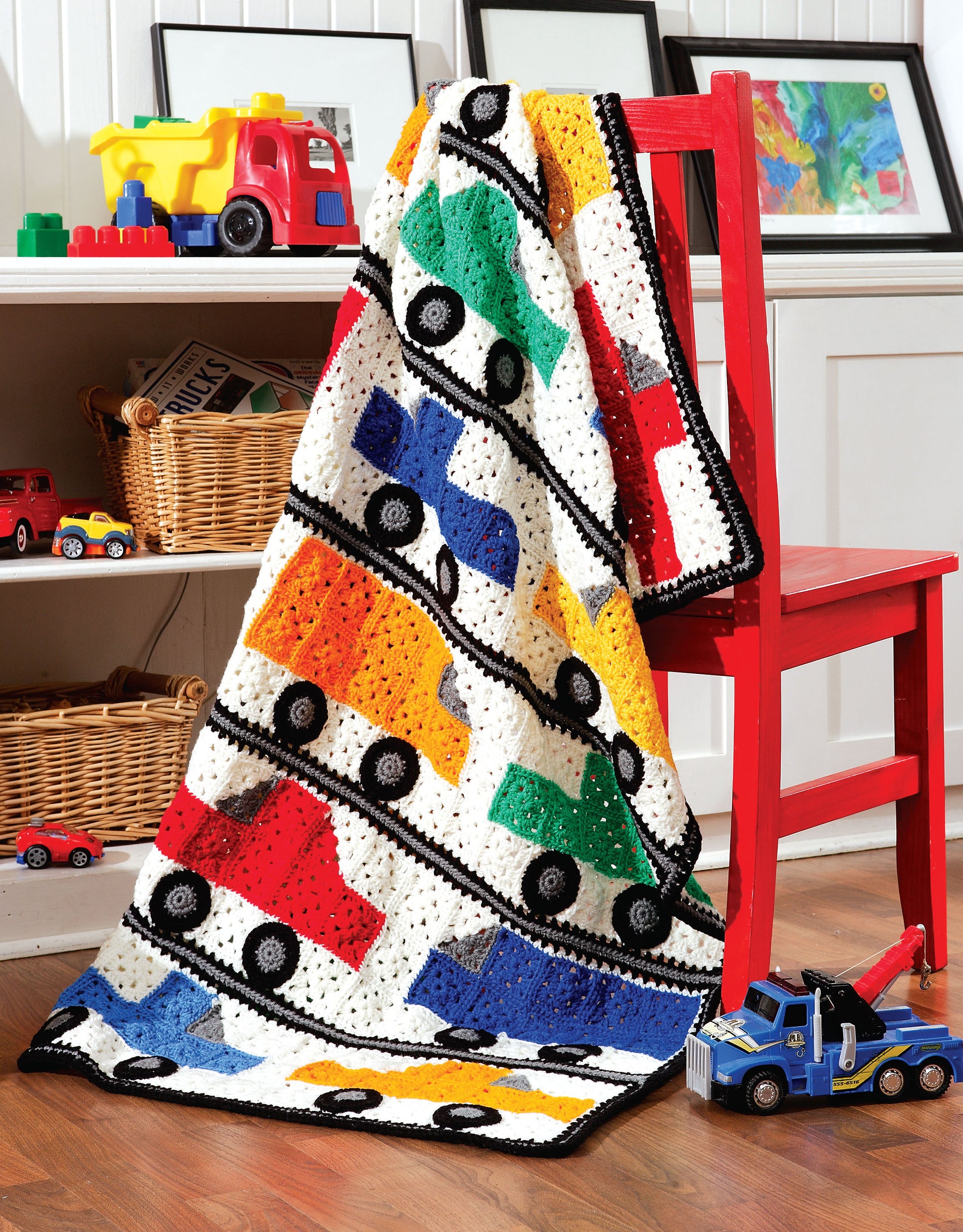 A vibrant crocheted blanket inspired by The Big Book of Baby Afghans from Leisure Arts, featuring rows of cars in red, yellow, green, and blue, is draped over a red chair in a playful child's room with toy trucks and cars on wooden floors.
