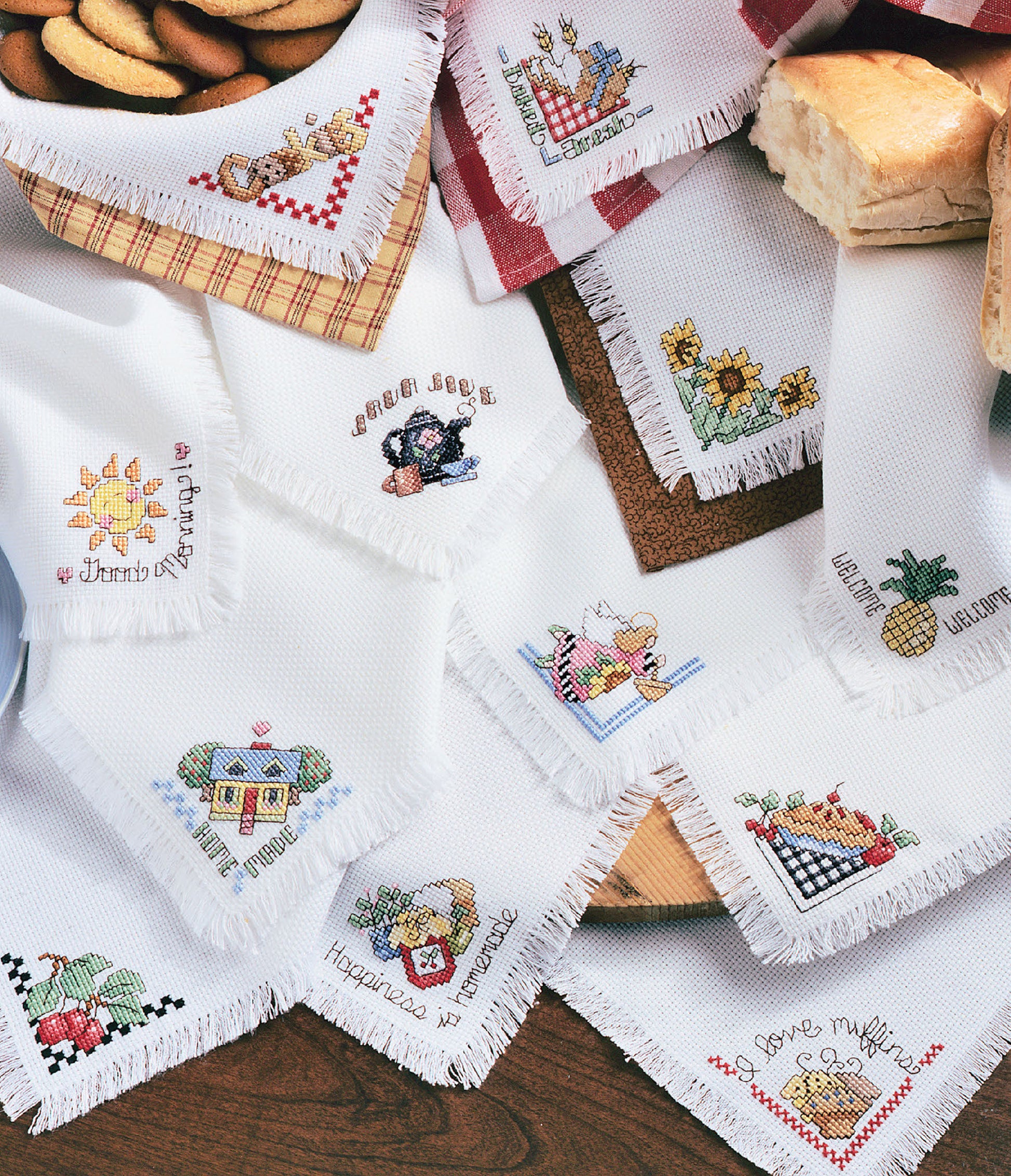 The Easy Bread Cloths Digital Download from Leisure Arts displays white napkins adorned with colorful cross stitch designs—flowers, fruit, houses, and hearts—arranged on a table with cookies and bread in the background.