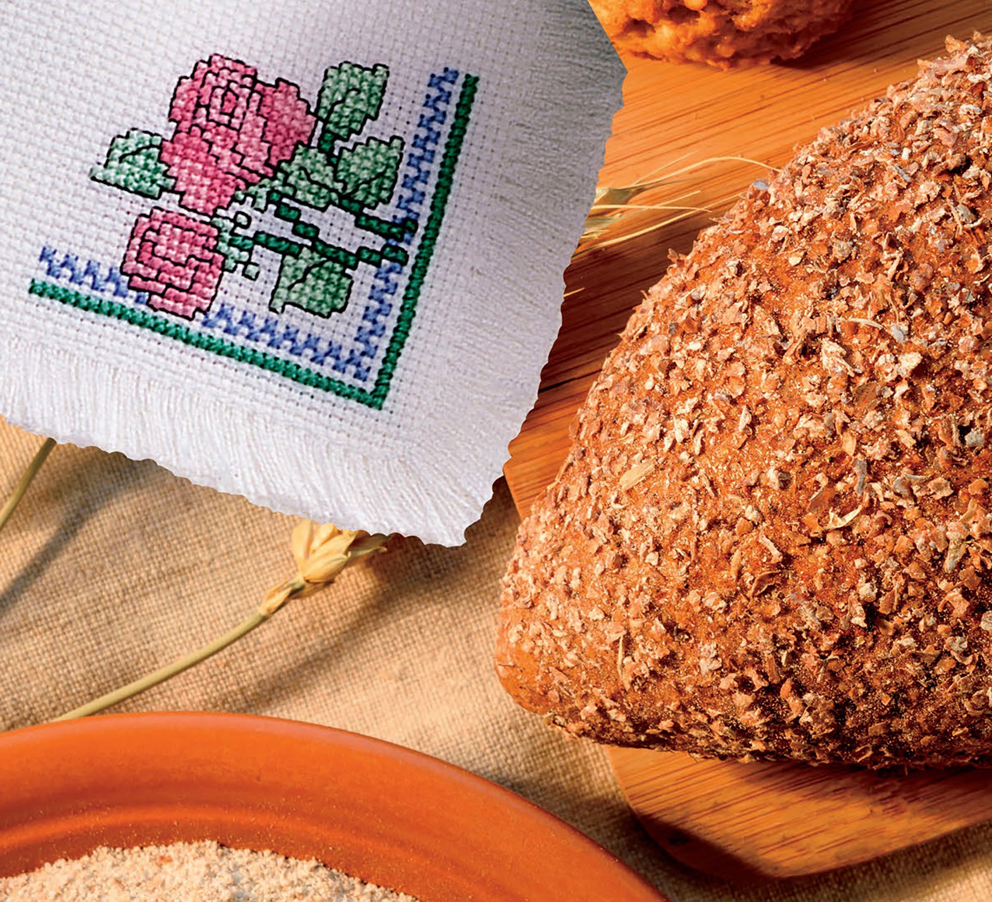 A close-up of whole grain bread rests on a wooden board beside the Easy Bread Cloths Digital Download from Leisure Arts, featuring cloth with pink and red rose embroidery. A bowl of flour and a wheat stalk appear in the background.