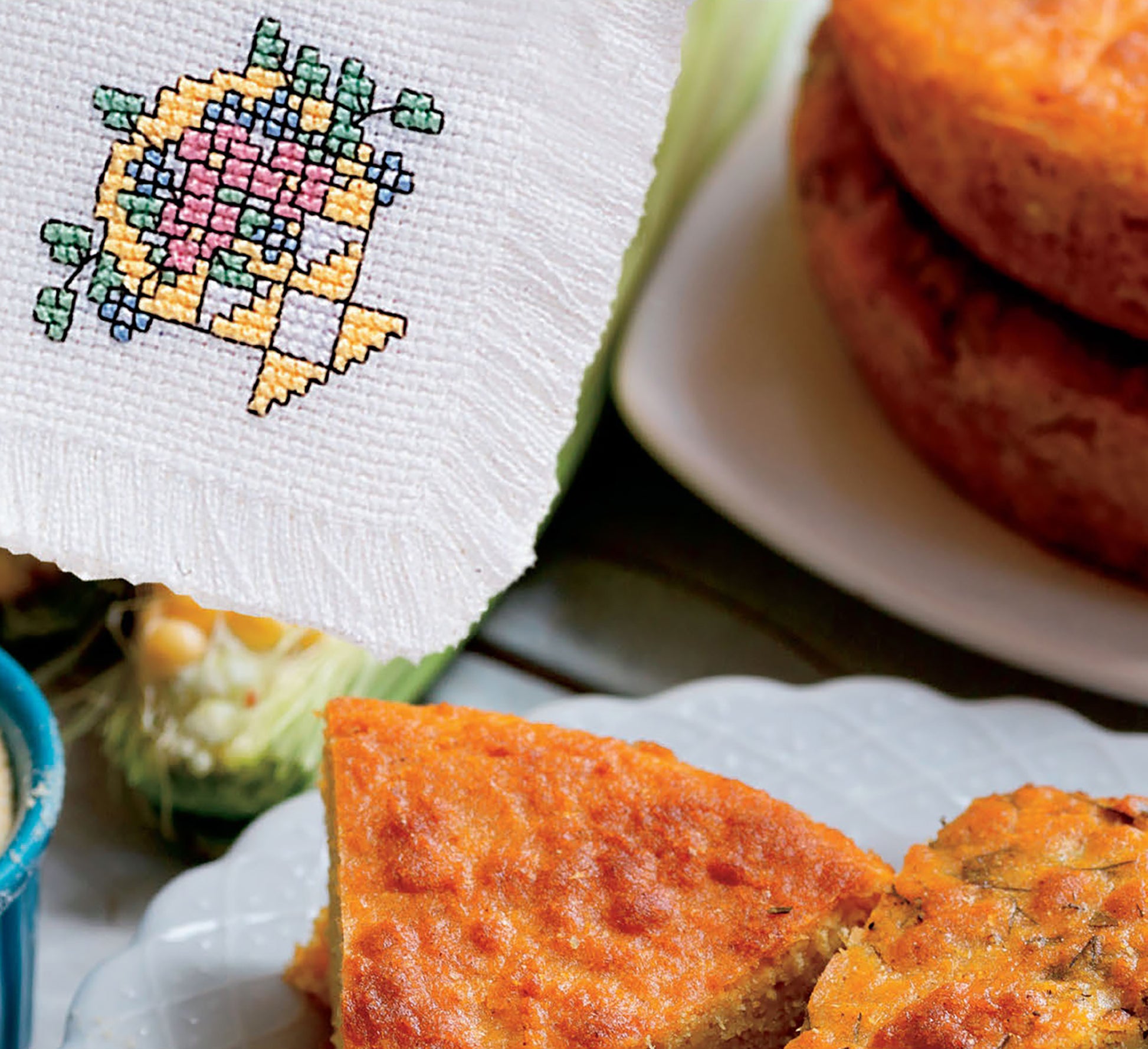 A close-up of cornbread on a white plate next to a napkin crafted with cross stitch designs from the Easy Bread Cloths Digital Download by Leisure Arts, featuring a yellow and green basket of pink flowers.
