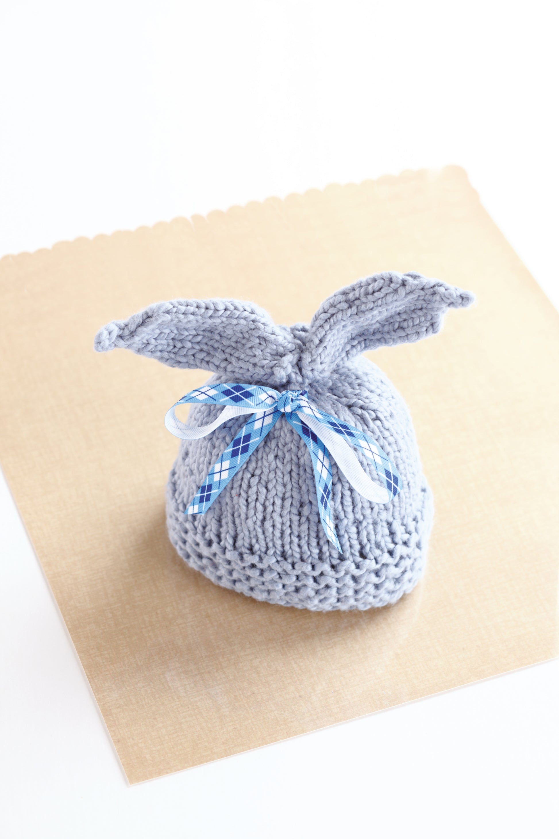 A small, hand-knitted gray pouch with tied ends and a blue and white checkered ribbon rests on a light brown square—great inspiration for "Warm Hats for Wee Noggins" by Leisure Arts or unique newborn knitting patterns.
