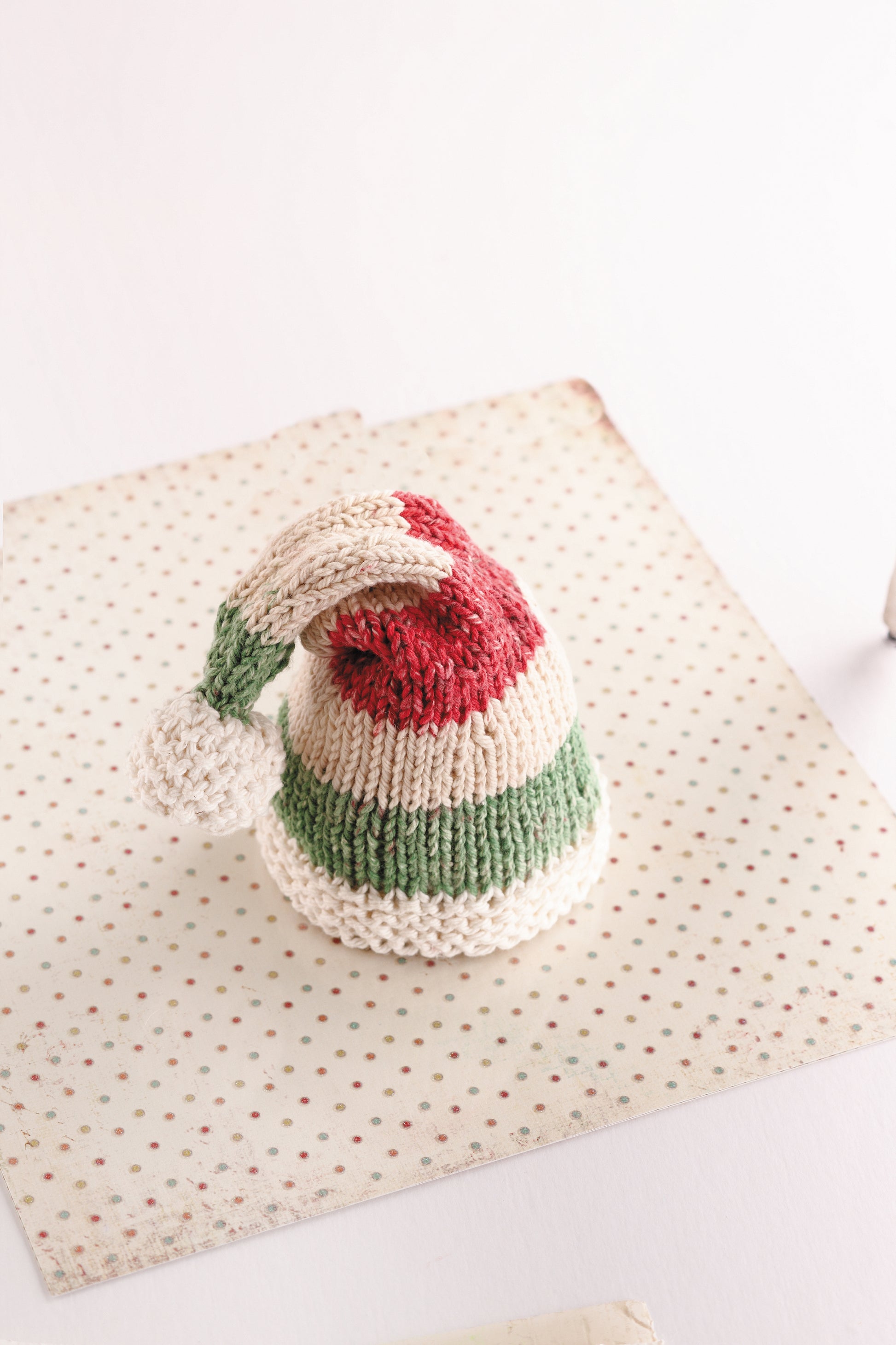 A colorful, striped knit hat with a white pom-pom from "Warm Hats for Wee Noggins" by Leisure Arts sits on a polka-dotted cloth—perfect inspiration for knit baby hats or preemie patterns.