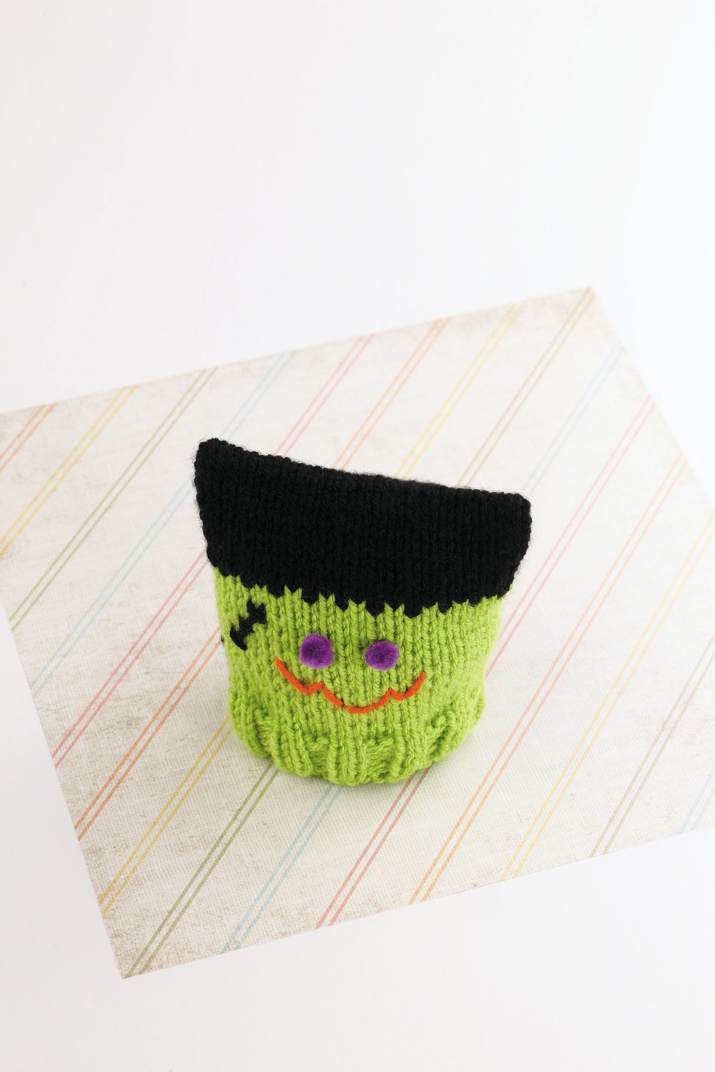 A green knitted cup cozy with a black top, purple eyes, orange smile, and scar—like a cute Frankenstein—rests on pastel stripes, inspiring baby projects from Warm Hats for Wee Noggins by Leisure Arts. Perfect for newborn knitting or preemie hat patterns.
