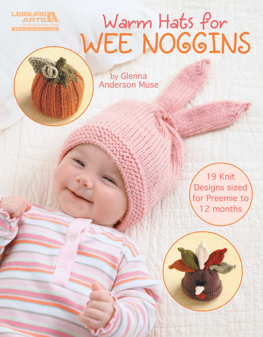 A smiling baby wears a pink striped outfit and a pink bunny-ear hat. Inset images show pumpkin and leaf hats. Text: “Warm Hats for Wee Noggins” by Leisure Arts—book of knit baby hat and preemie hat patterns.