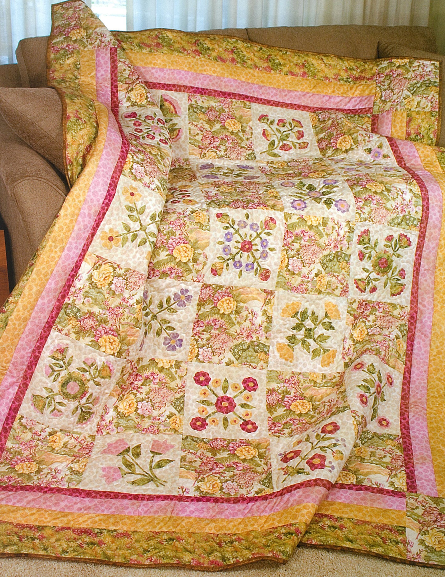The "100 Any-Size Applique Blocks" quilt by Leisure Arts, featuring pink, yellow, and green floral and leaf patterns, drapes over a brown couch to add a cozy and decorative touch to the room.