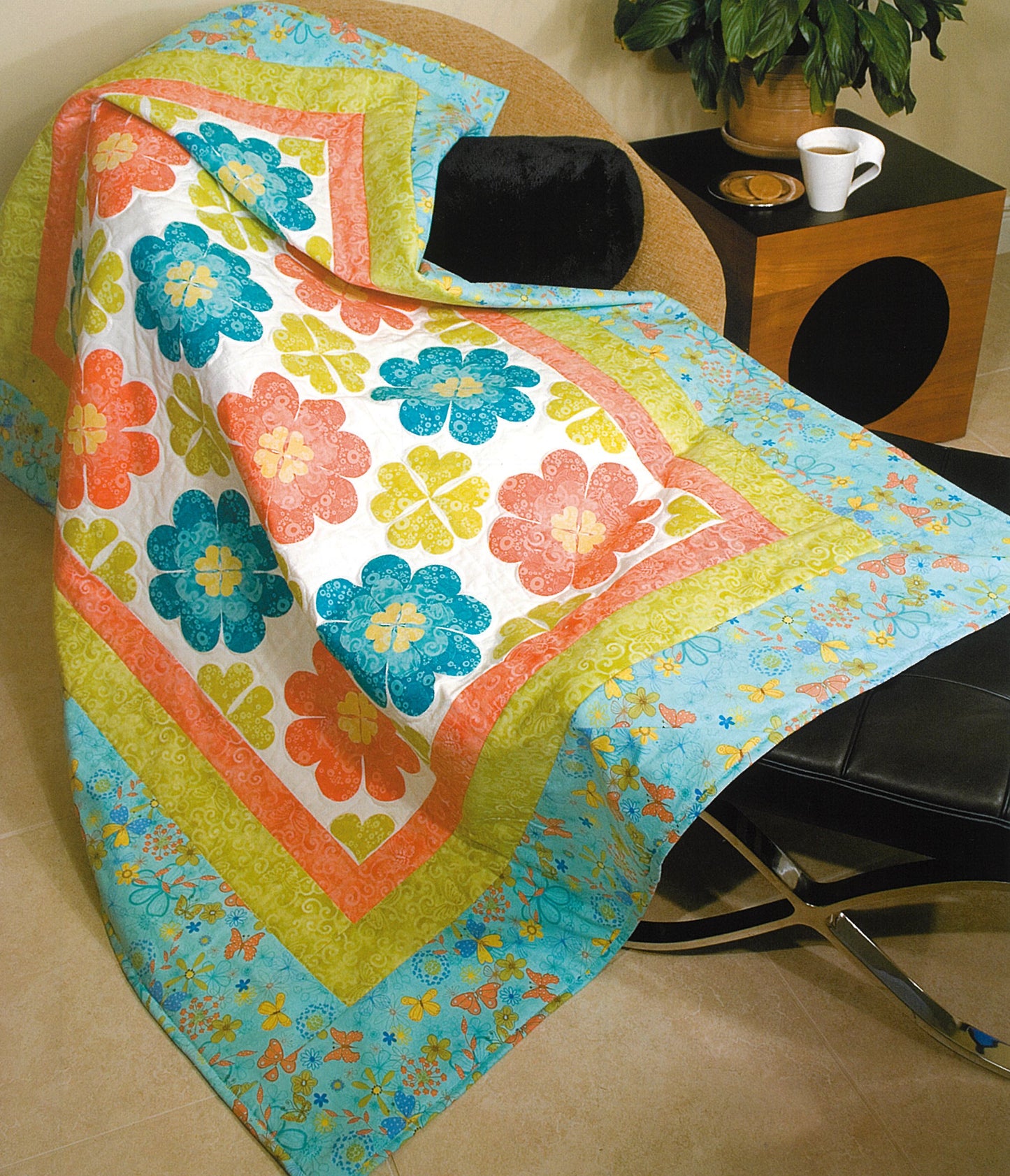 A quilt made with blue, orange, green, and yellow floral patterns using Leisure Arts' 100 Any-Size Applique Blocks is draped over a chair by a wooden table holding a plant and cup.