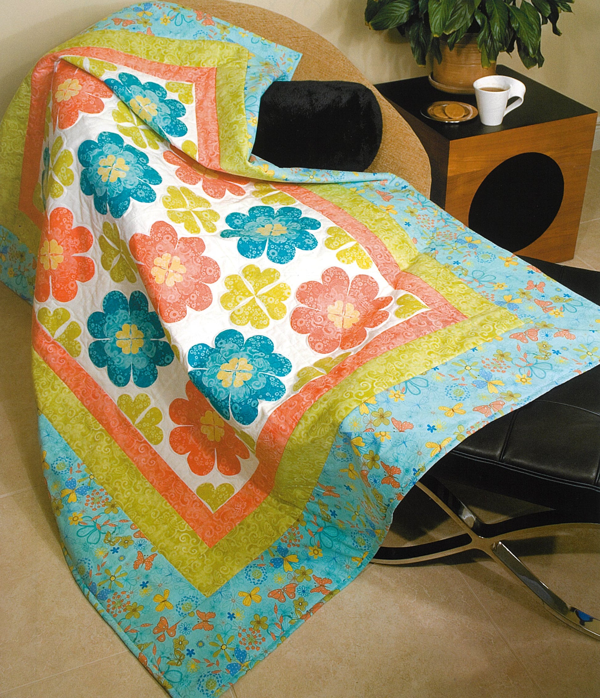 A quilt made with blue, orange, green, and yellow floral patterns using Leisure Arts' 100 Any-Size Applique Blocks is draped over a chair by a wooden table holding a plant and cup.
