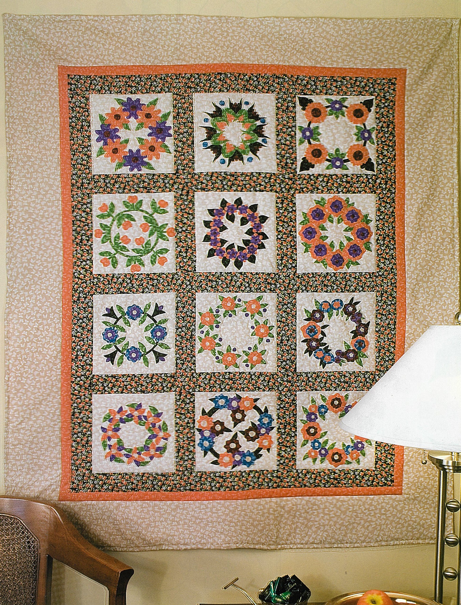 A vibrant quilt made from Leisure Arts' 100 Any-Size Applique Blocks hangs on the wall, each of its twelve squares showcasing unique floral patterns. The pink and black floral border complements a lamp and part of a chair in the foreground.