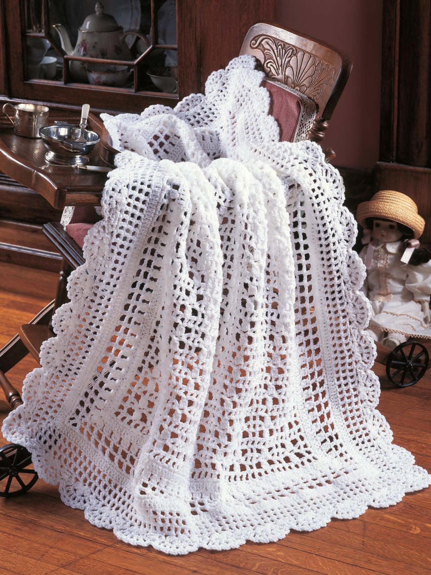 A Leisure Arts "Heirloom Afghans for Baby" white crocheted blanket with scalloped edges drapes over a vintage stroller; behind it, a wooden cabinet holds teapots and a porcelain doll in a hat sits on the floor.