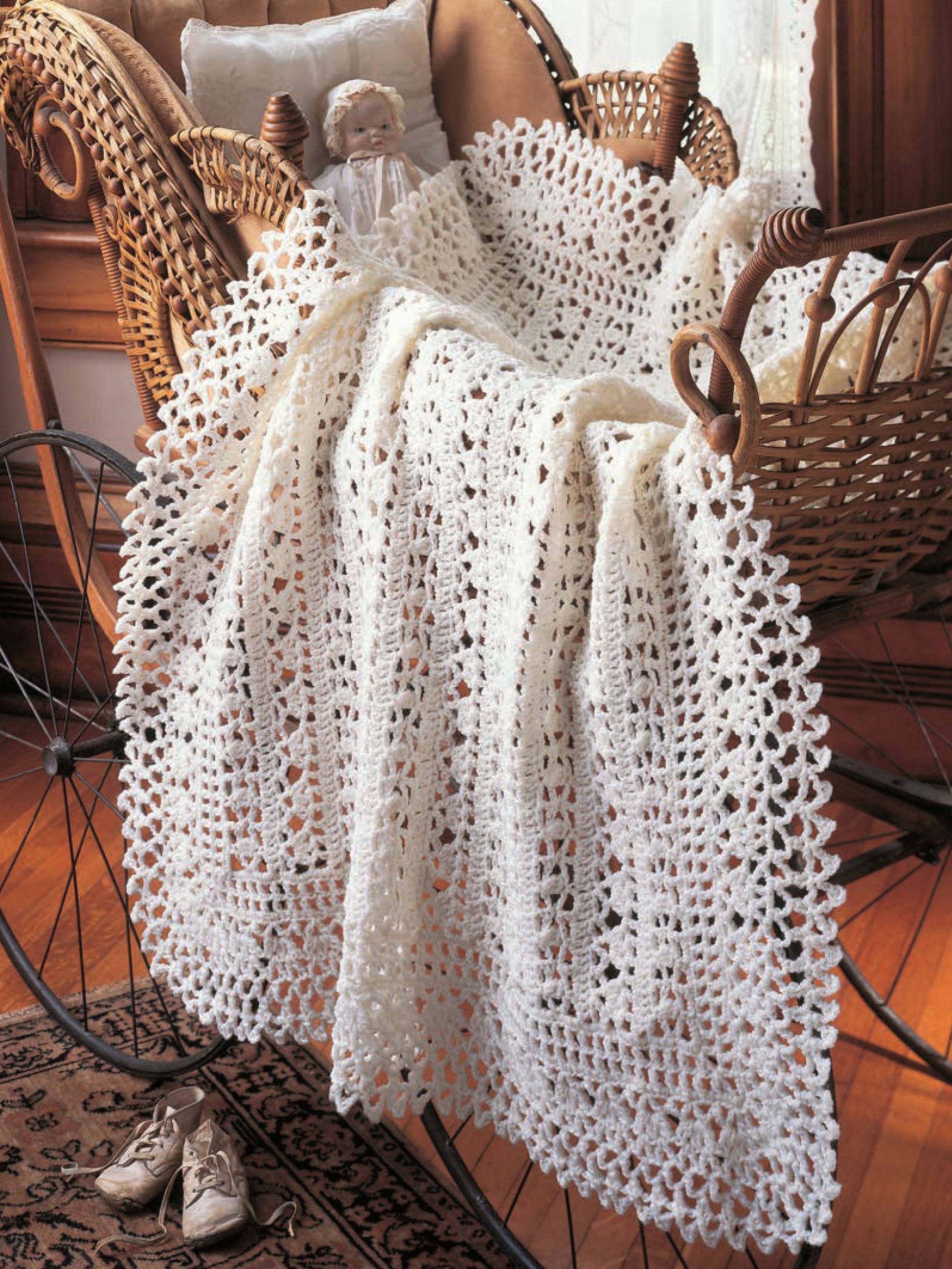 A vintage wicker baby carriage holds a doll and is draped with a white crocheted blanket made from Heirloom Afghans for Baby by Leisure Arts, with baby shoes placed on the floor nearby.