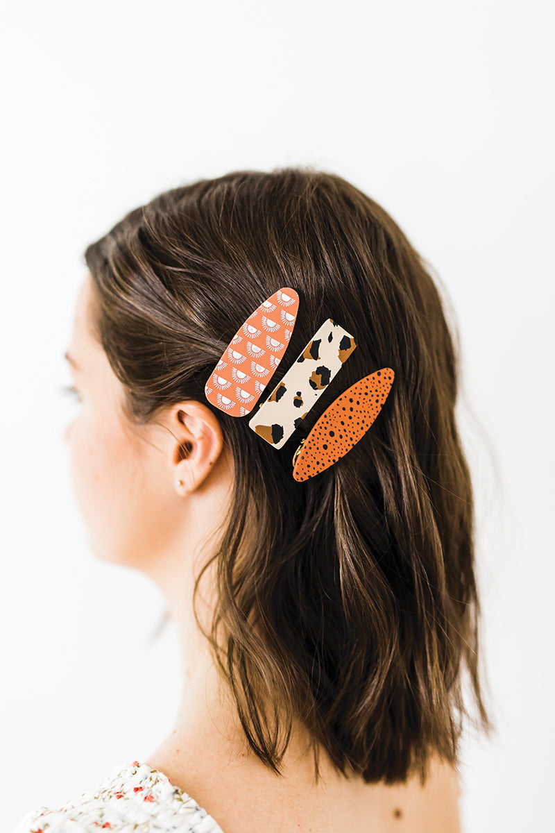 A woman with shoulder-length brown hair wears three large Polymer Clay Hair Clips by C&T Publishing in various patterns: pink with white semicircles, beige with black and brown spots, and orange with small black dots.