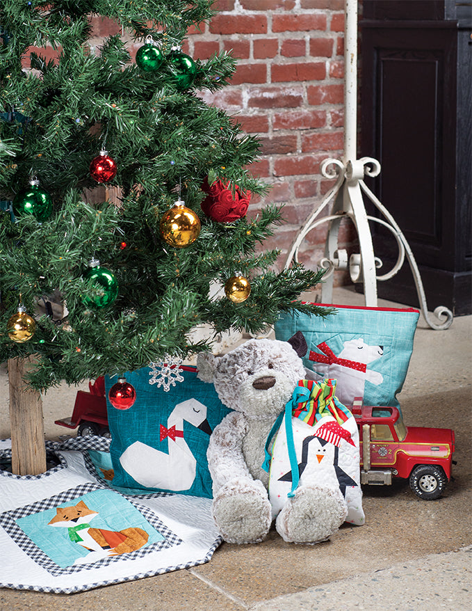 A decorated Christmas tree stands next to a gray stuffed bear, penguin-themed gift bags, a red toy truck, and quilted animal-themed creations from *Sew a Winter Woodland Christmas* by C&T Publishing, inspired by paper pieced woodland blocks. The background features a brick wall and part of a metal stand, perfectly capturing the cozy spirit of holiday home decor sewing.