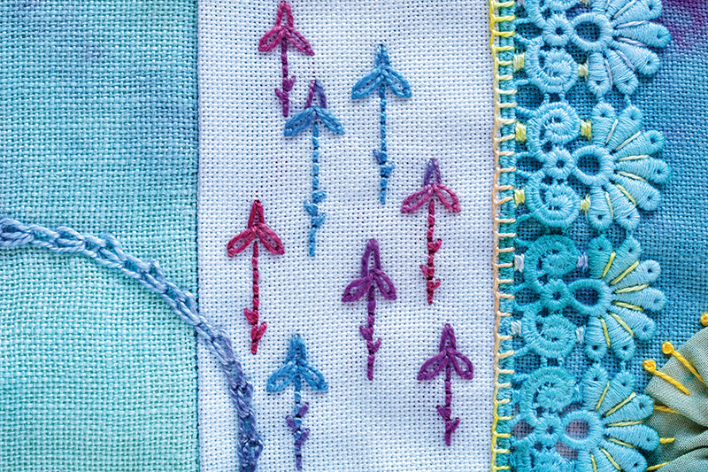 Close-up from C&T Publishing’s "Creative Stitches for Contemporary Embroidery, Volume 2" features pink and blue stitched arrows on white fabric, bordered by blue patterned fabric, lace trim, and decorative embroidery—perfect for crazy quilting.