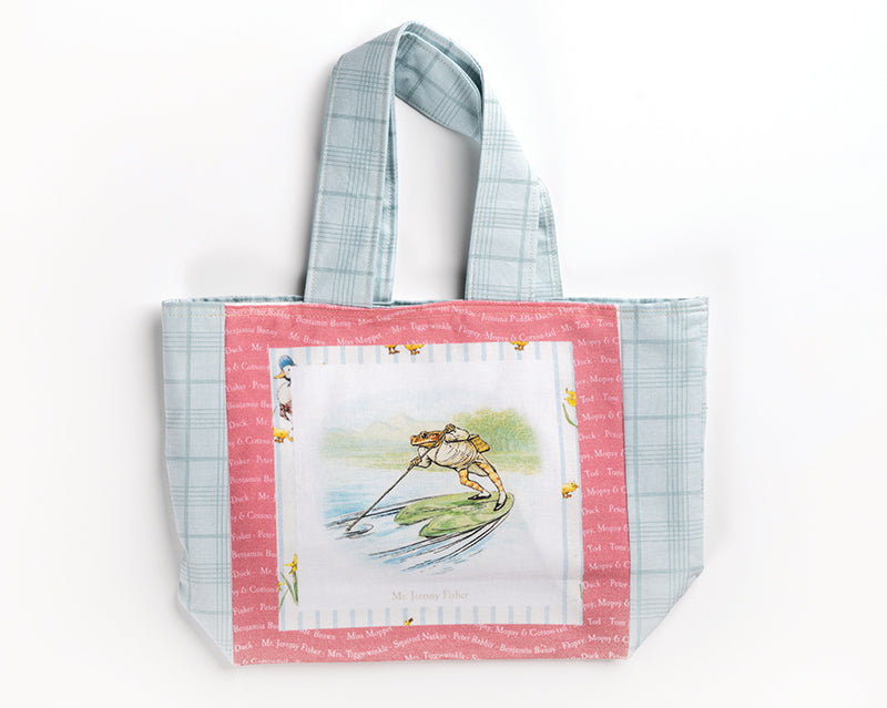 The Quilt Panel Magic tote bag from C&T Publishing features blue plaid handles, pink trim with white text, and a frog fishing by a pond. "Mr. Jeremy Fisher" appears below the art—ideal for carrying panel quilts or instructions.