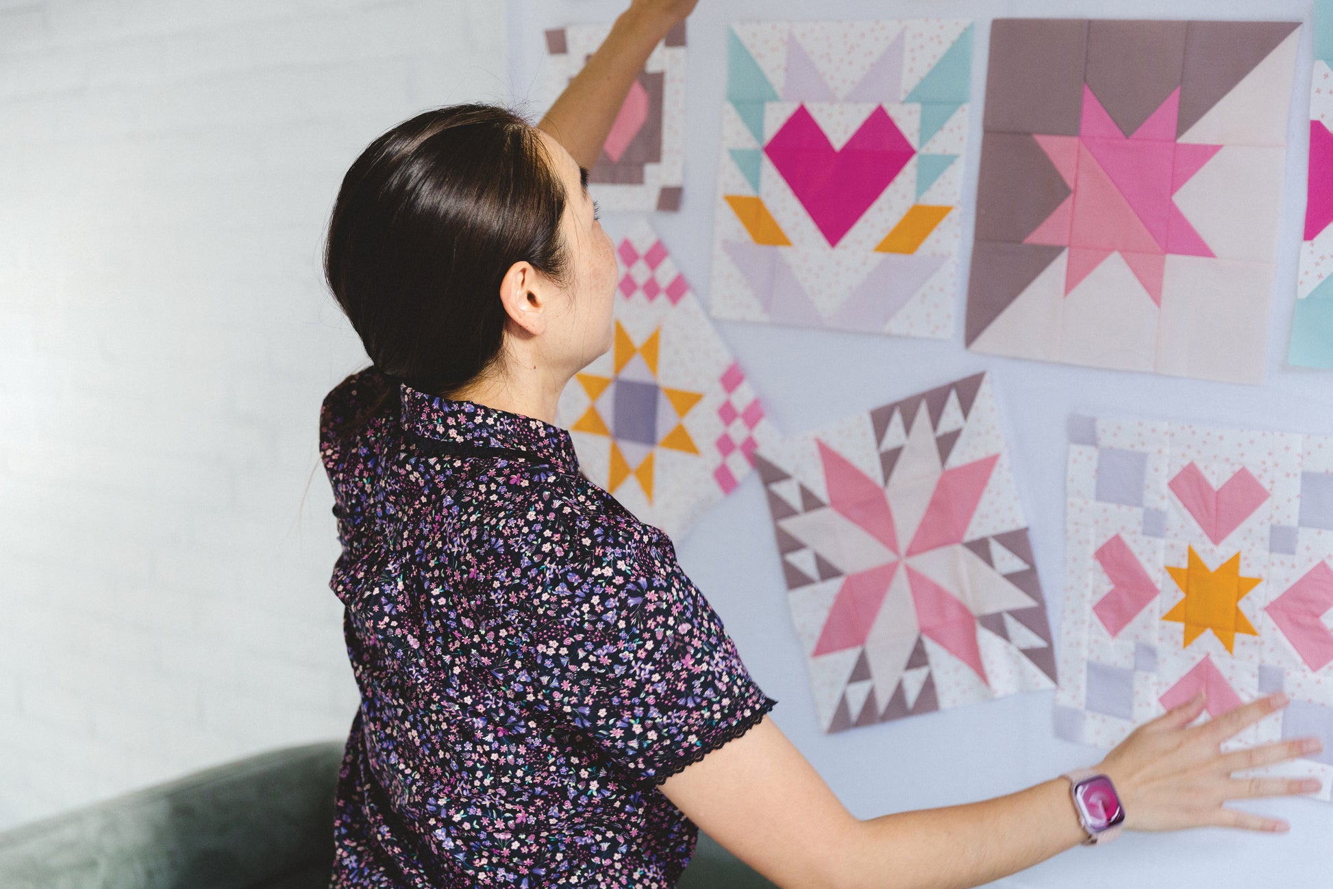 A person with dark hair arranges Patchwork Playground quilt blocks by Stash Books on a white wall. These modern quilts display geometric patterns and heart shapes in colorful pink, purple, yellow, and white patchwork.