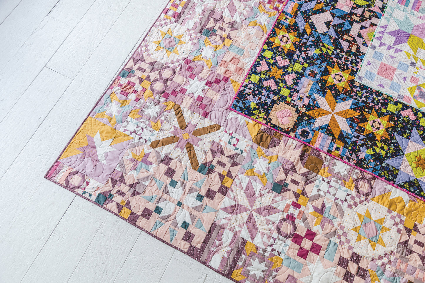A close-up of "Patchwork Playground" by Stash Books—a vibrant patchwork quilt with intricate geometric and star blocks in pink, yellow, purple, and blue—displayed on a white wooden floor.