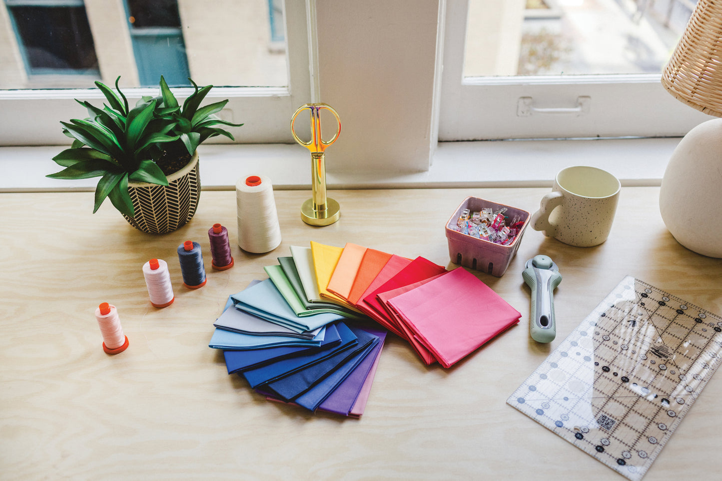 The Patchwork Playground by Stash Books features colorful fabric squares in a rainbow fan, spools of thread, rotary cutter, clear ruler, scissors, sewing clips in a basket—an ideal setup for modern quilts.