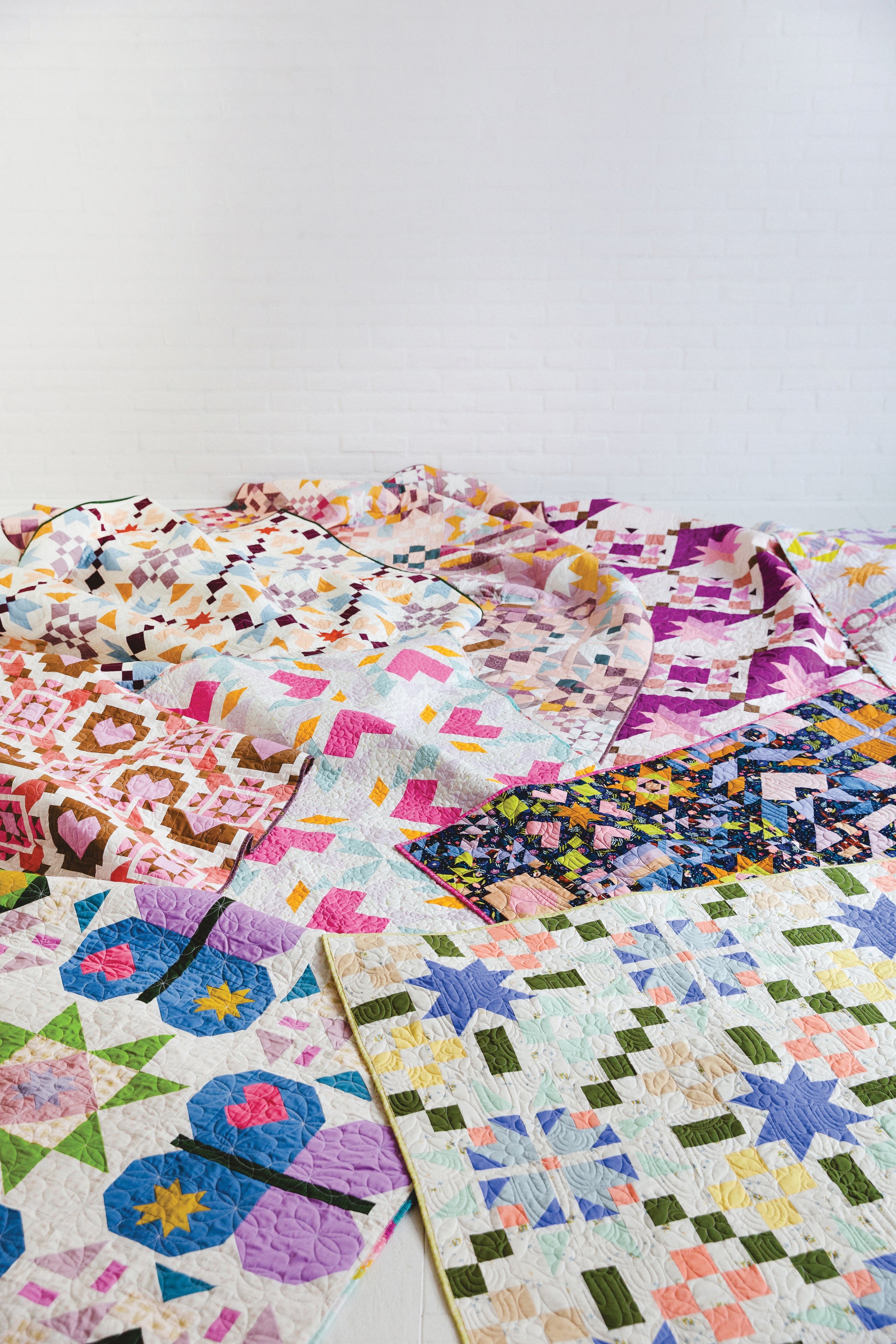 Patchwork Playground by Stash Books features vibrant quilts with modern geometric and floral patterns, beautifully displayed on a white floor in front of a white brick wall.