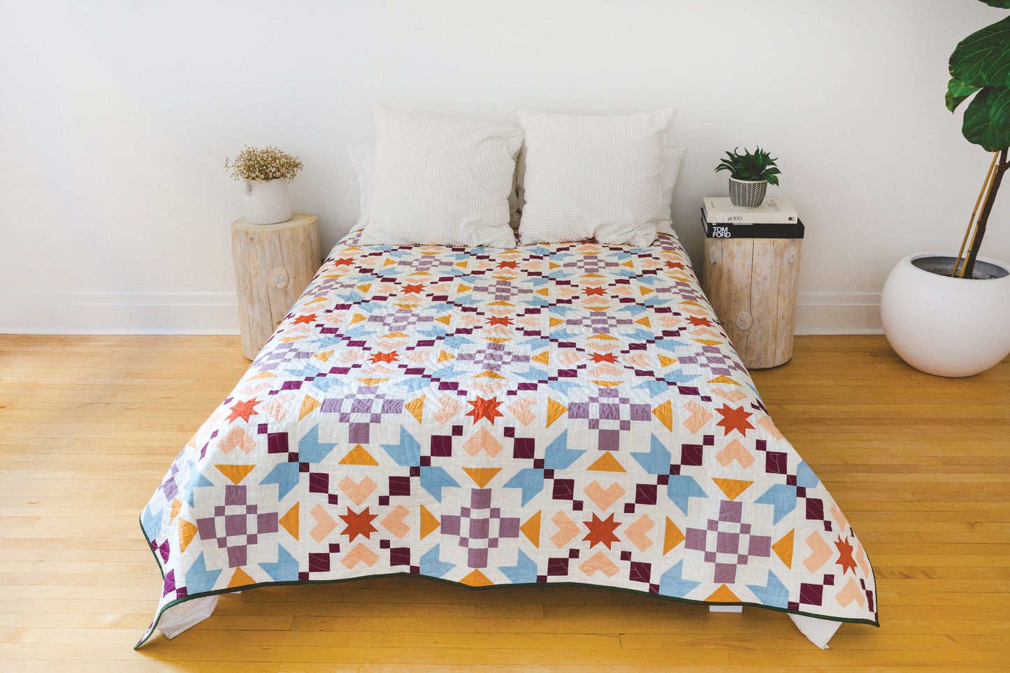 A neatly made bed with two white pillows and the Patchwork Playground quilt by Stash Books, boasting bold modern blocks, sits between two wooden nightstands—one with a plant, the other with books—on a light wood floor.