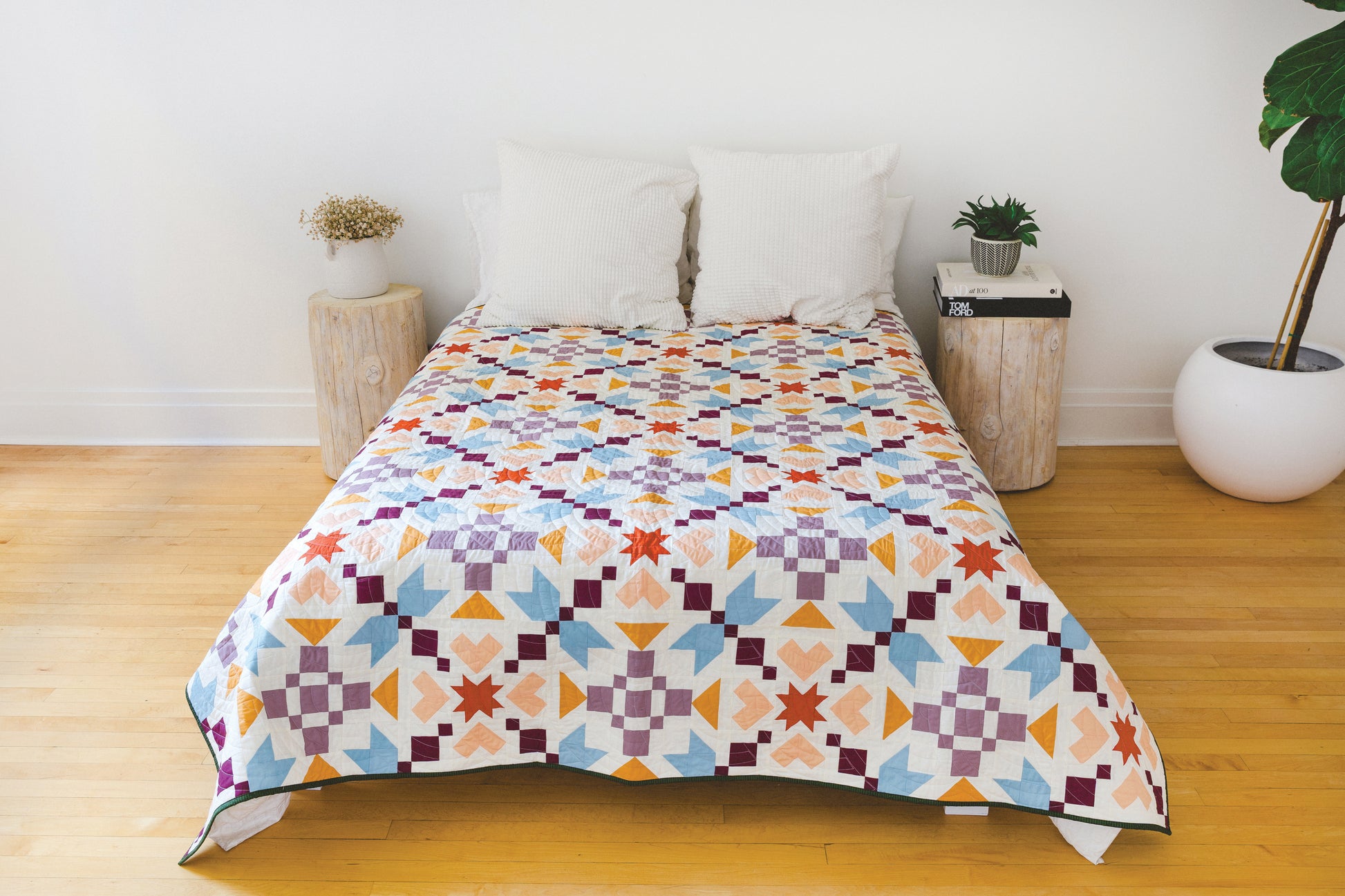 A neatly made bed with two white pillows and the Patchwork Playground quilt by Stash Books, boasting bold modern blocks, sits between two wooden nightstands—one with a plant, the other with books—on a light wood floor.