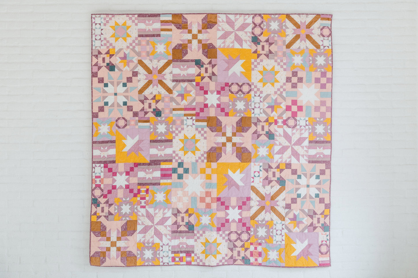 Patchwork Playground by Stash Books features vibrant pink, yellow, white, and purple geometric star, square, and cross blocks. Displayed against a white brick wall—perfect for modern quilt enthusiasts.