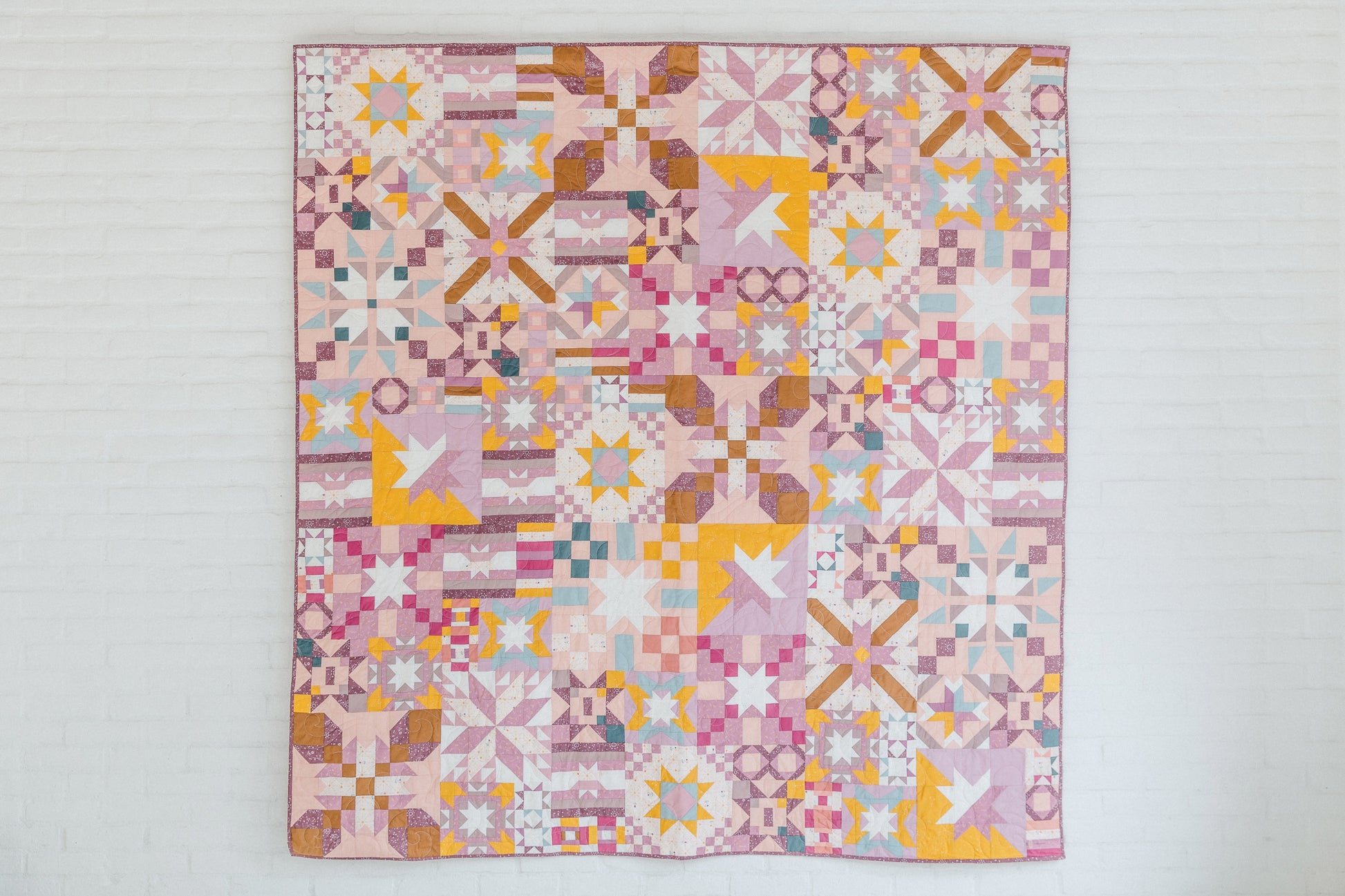 Patchwork Playground by Stash Books features vibrant pink, yellow, white, and purple geometric star, square, and cross blocks. Displayed against a white brick wall—perfect for modern quilt enthusiasts.