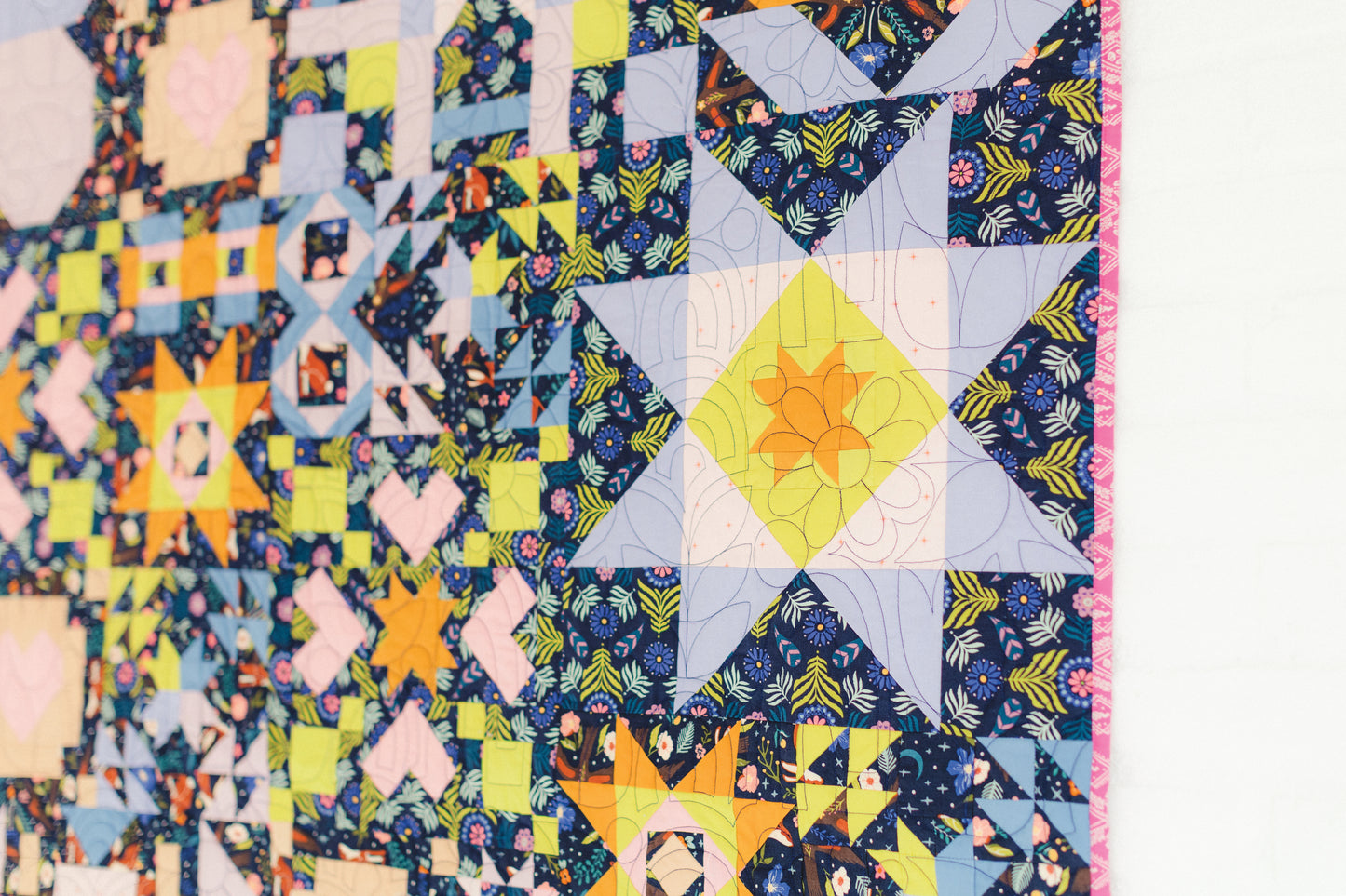 “Patchwork Playground” by Stash Books hangs on a white wall, featuring vibrant star and geometric quilt blocks in blue, yellow, pink, and green with intricate fabric designs and detailed stitching.