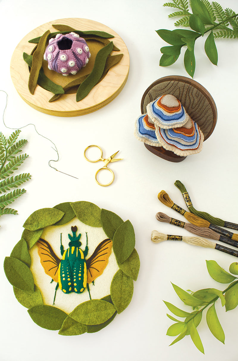 A flat lay of embroidered felt art from "Felt & Thread" by Stash Books, featuring a green beetle with yellow wings, purple sea urchin, layered mushrooms, embroidery floss, scissors, and green leaves on a white background.