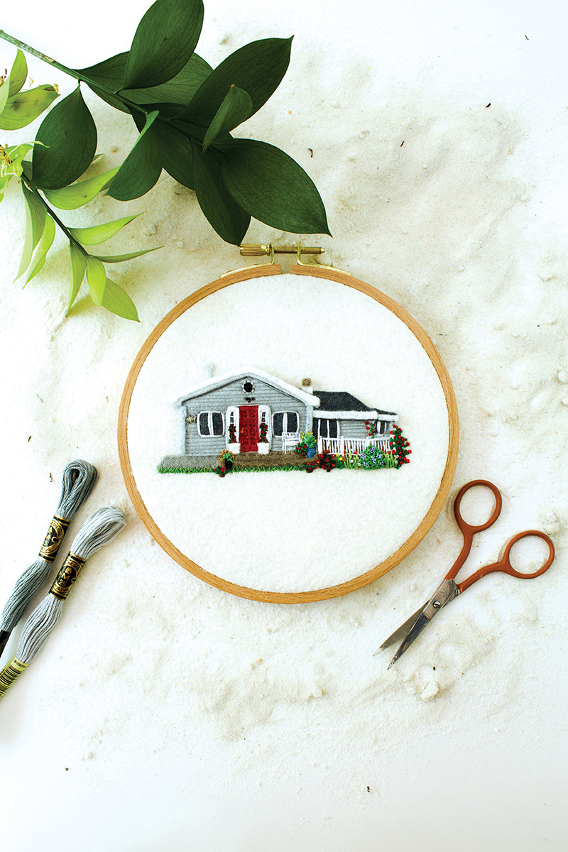 The Felt & Thread embroidery kit from Stash Books features a stitched gray house with a red door surrounded by greenery, plus threads for thread painting, small scissors, and leafy branches arranged on a white surface.