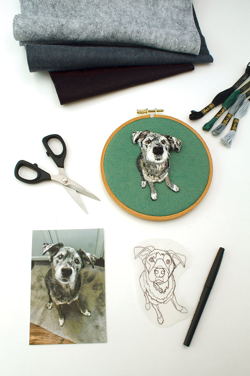 A flat lay of Felt & Thread by Stash Books displays fabric, thread, scissors, a pen, a dog photo, a transparent sketch of a dog, and an embroidery hoop with a thread-painted dog portrait.