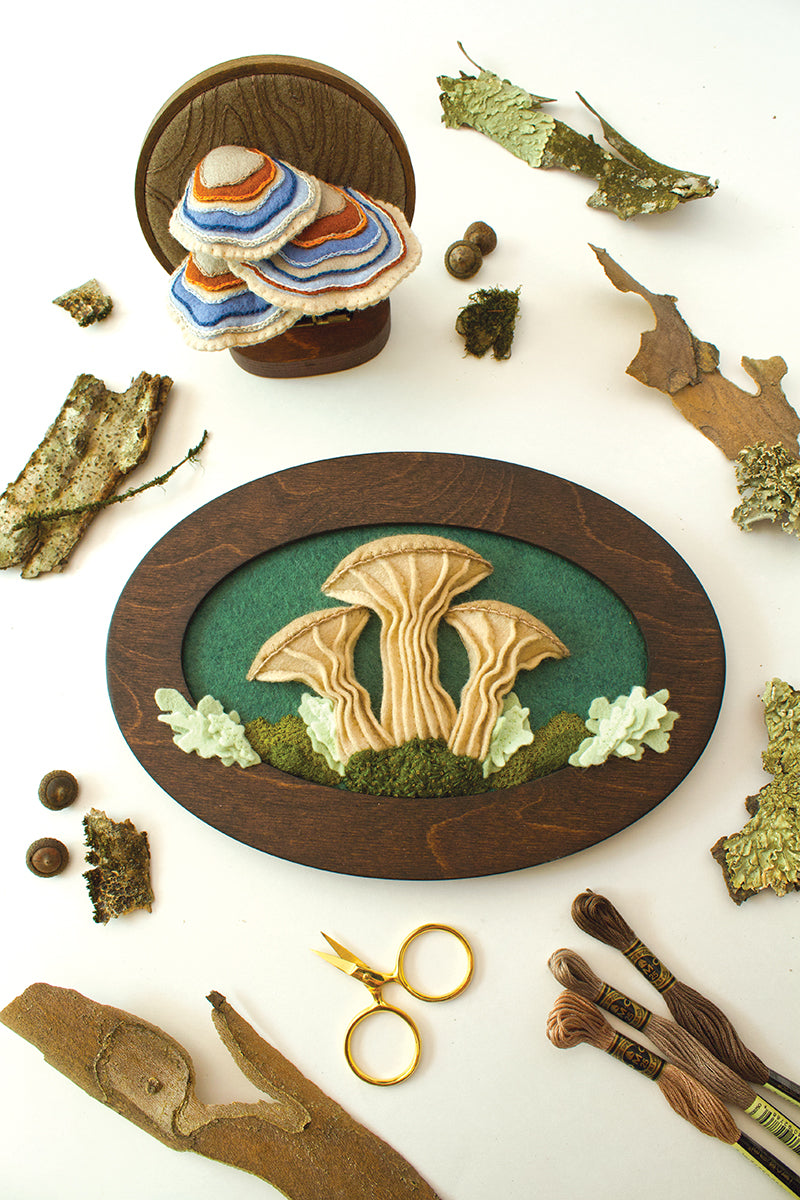 A flat lay from Stash Books’ "Felt & Thread" shows embroidered mushroom art in wooden frames with felt appliqué, surrounded by bark, acorns, embroidery floss, gold scissors, and sewing needles on a white background.