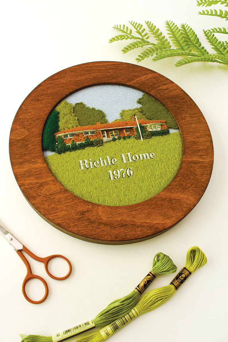 A round wooden embroidery hoop from Stash Books’ Felt & Thread displays a stitched house, green lawn, blue sky, and “Richie Home 1976.” Nearby are green embroidery floss, scissors, and a fern.