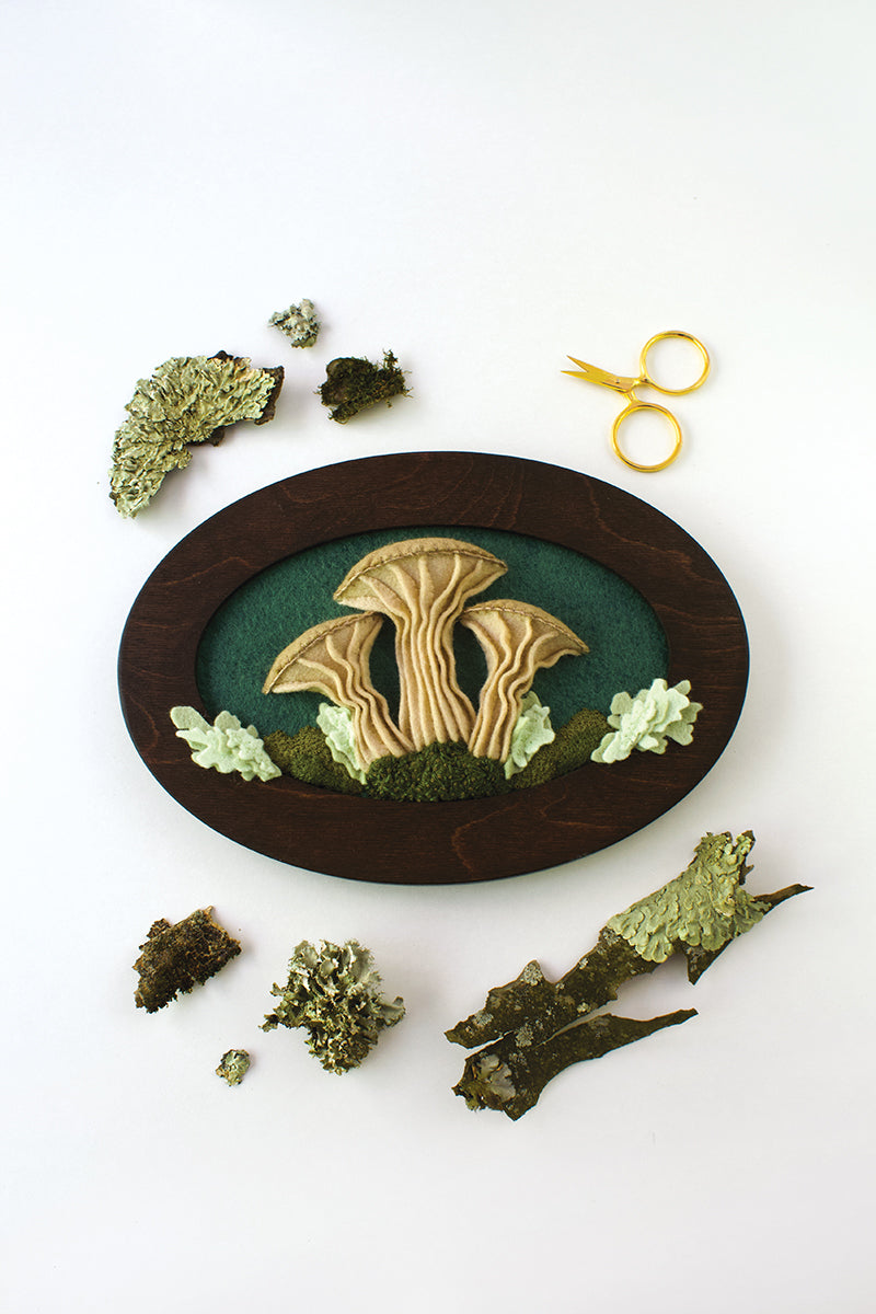 The "Felt & Thread" artwork by Stash Books features embroidered mushrooms in an oval wooden frame, displayed on a white surface with moss, lichen, and small gold scissors.