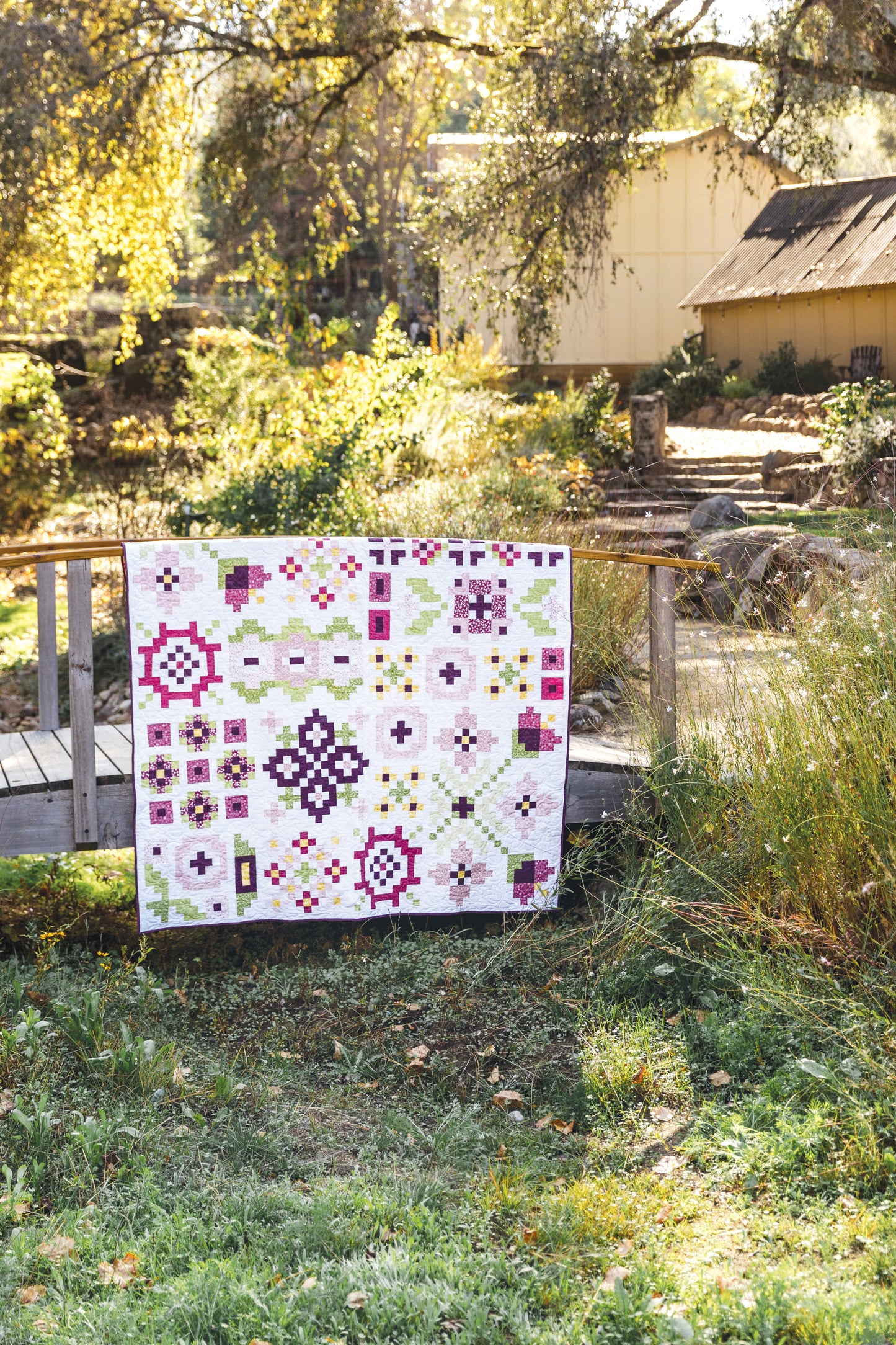 The Garden Dalliance Sampler by C&T Publishing—a vibrant quilt with geometric designs—hangs over a wooden bridge in a sunlit garden with tall grass, trees, and a rustic building in the background.