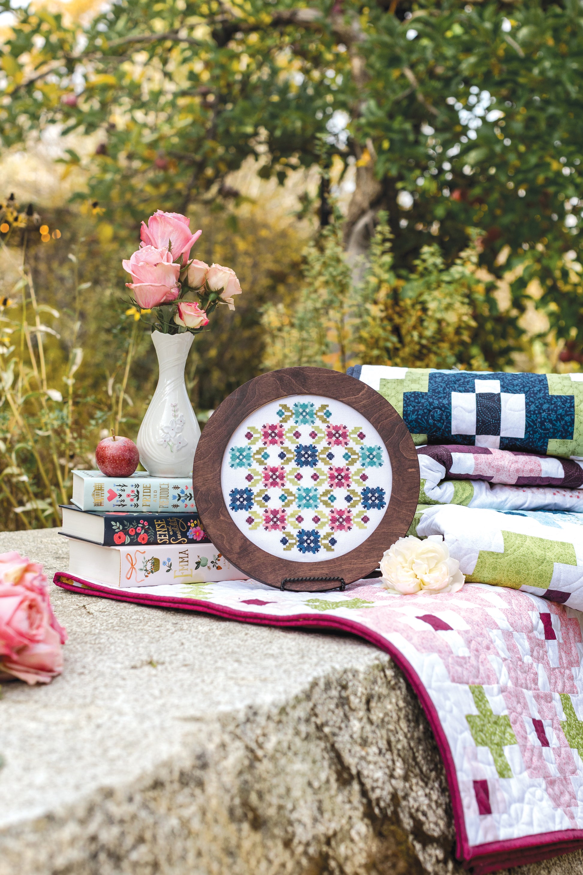 The Garden Dalliance Sampler by C&T Publishing, in a wooden frame, rests on a stone table outdoors with folded quilts, a vase of pink roses, books, an apple, and a white flower among lush greenery.