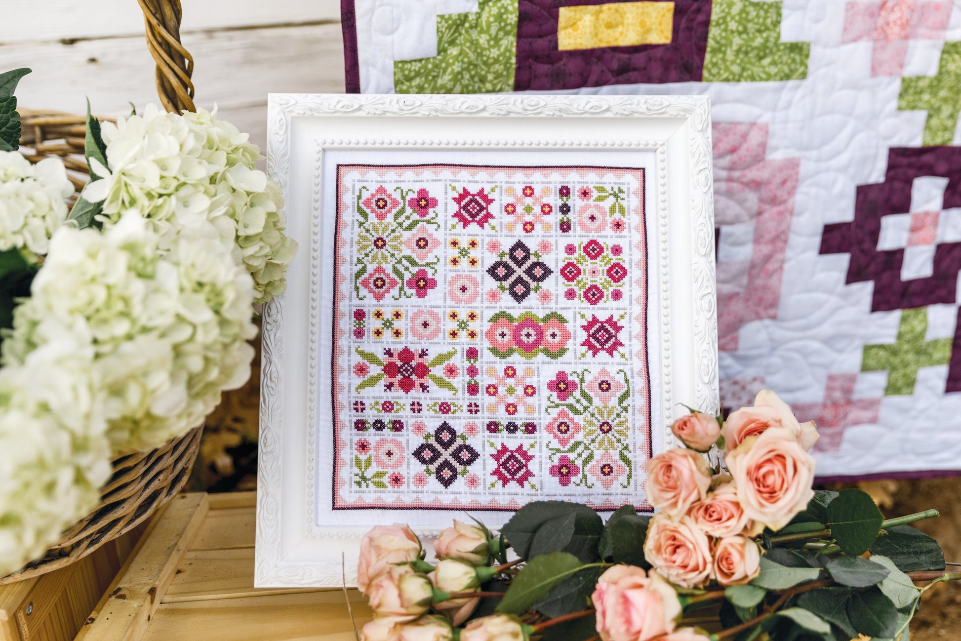 The Garden Dalliance Sampler by C&T Publishing, a framed cross-stitch with vibrant geometric and floral designs, is displayed on wood among white hydrangeas and peach roses, with a quilt in the background.