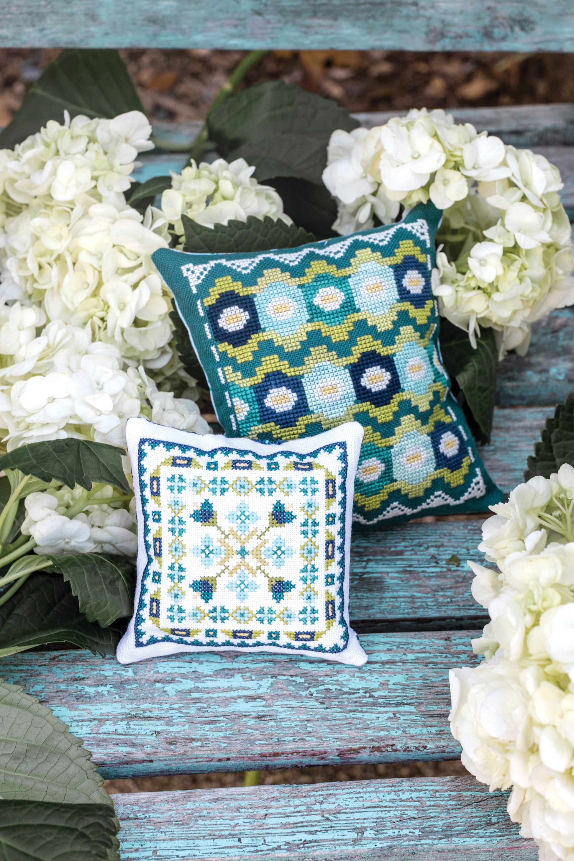 Two small, colorful pillows made with the Garden Dalliance Sampler by C&T Publishing rest on a weathered turquoise bench, surrounded by clusters of white hydrangea flowers and green leaves.