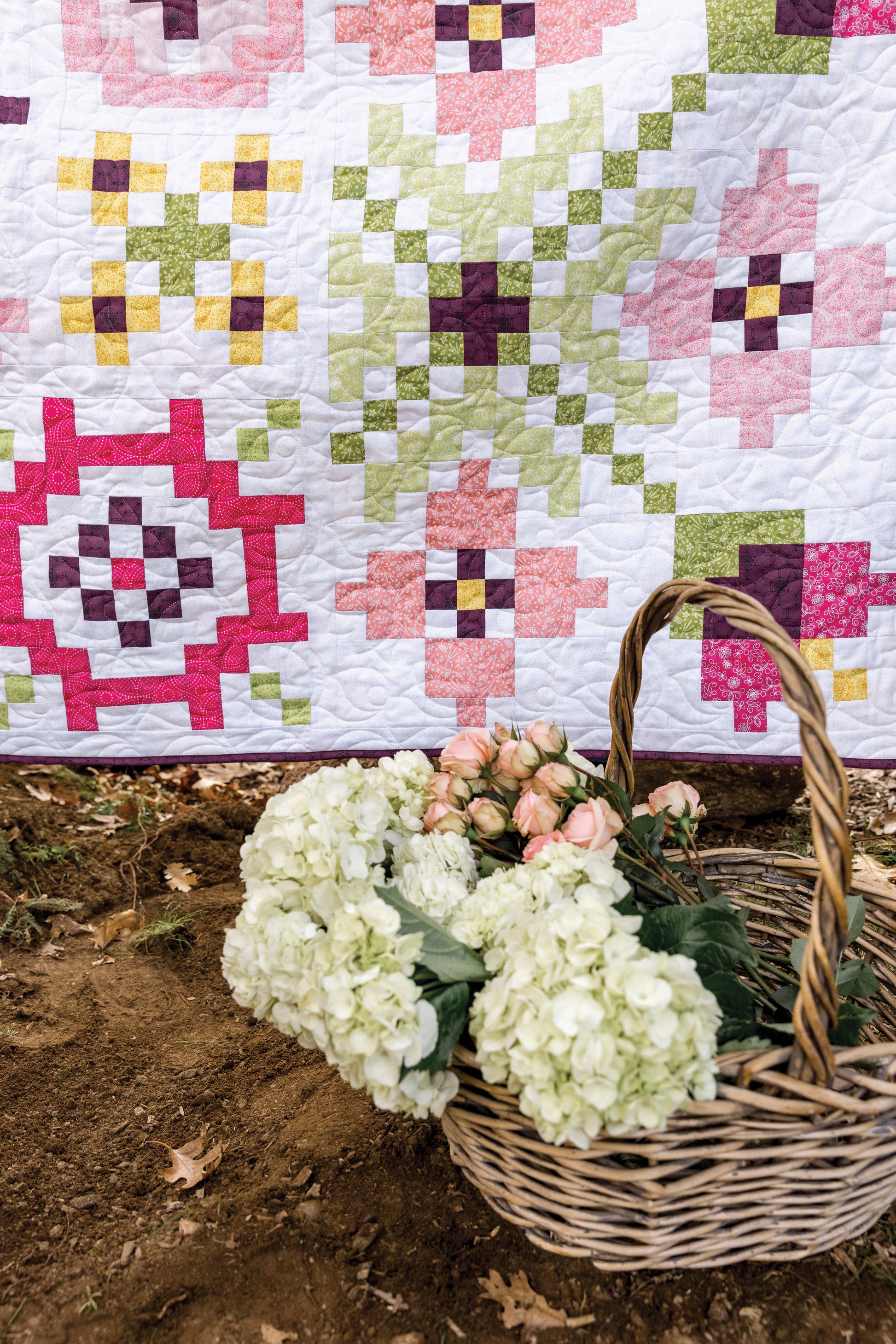 The C&T Publishing Garden Dalliance Sampler, with its pastel florals, sits before a colorful patchwork quilt—offering perfect inspiration for your next project and serving as a charming backdrop.