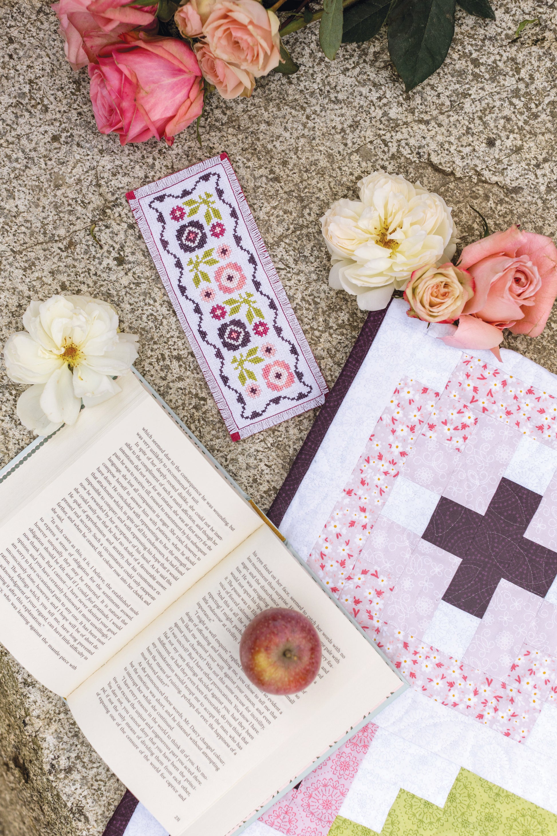 An open book with a red apple rests on stone next to quilt projects, the Garden Dalliance Sampler floral bookmark by C&T Publishing, and pink and white roses.