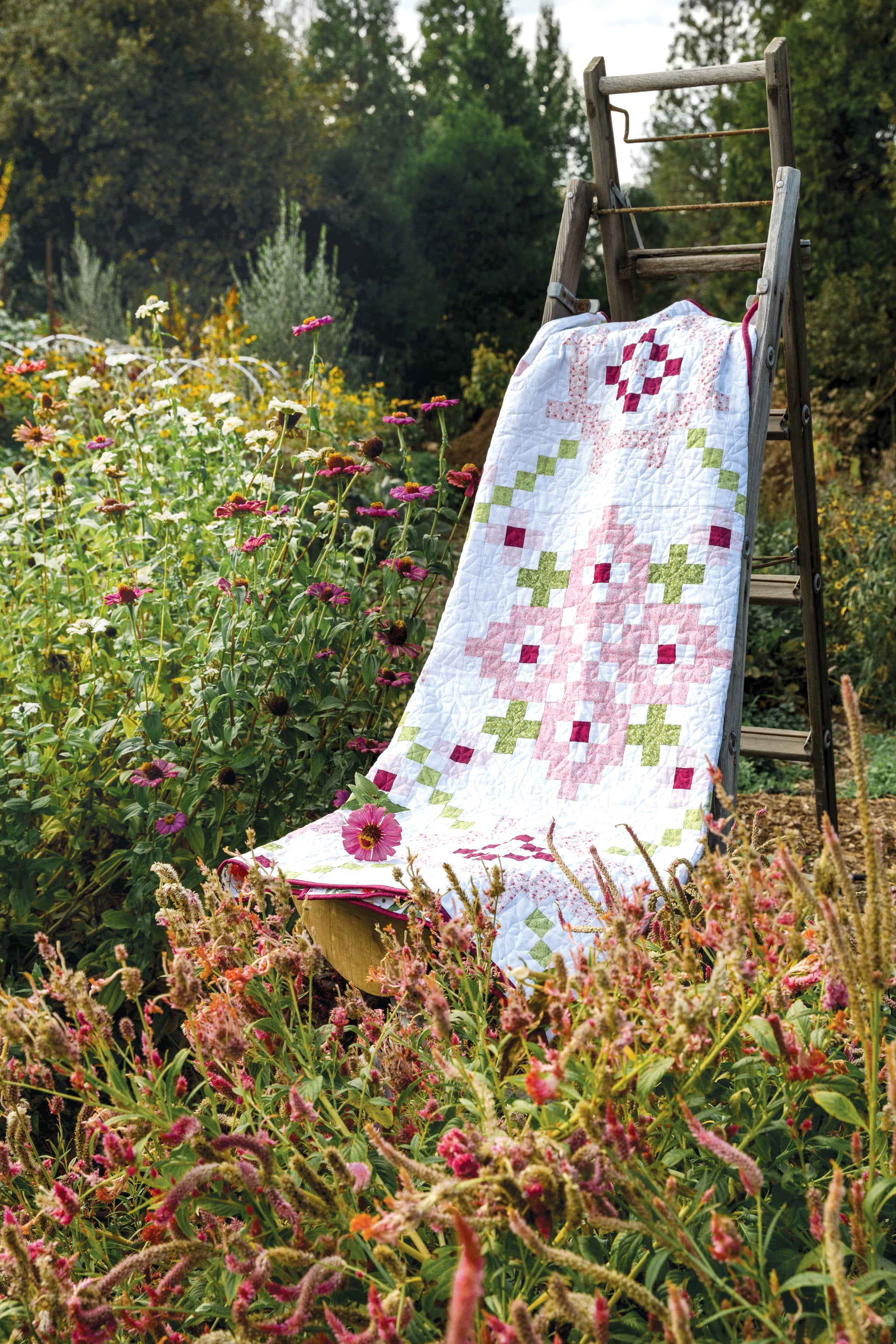 The Garden Dalliance Sampler by C&T Publishing, featuring pink, green, and white patterns ideal for quilting projects, is displayed on a wooden ladder amidst blooming flowers and lush greenery in a vibrant garden.