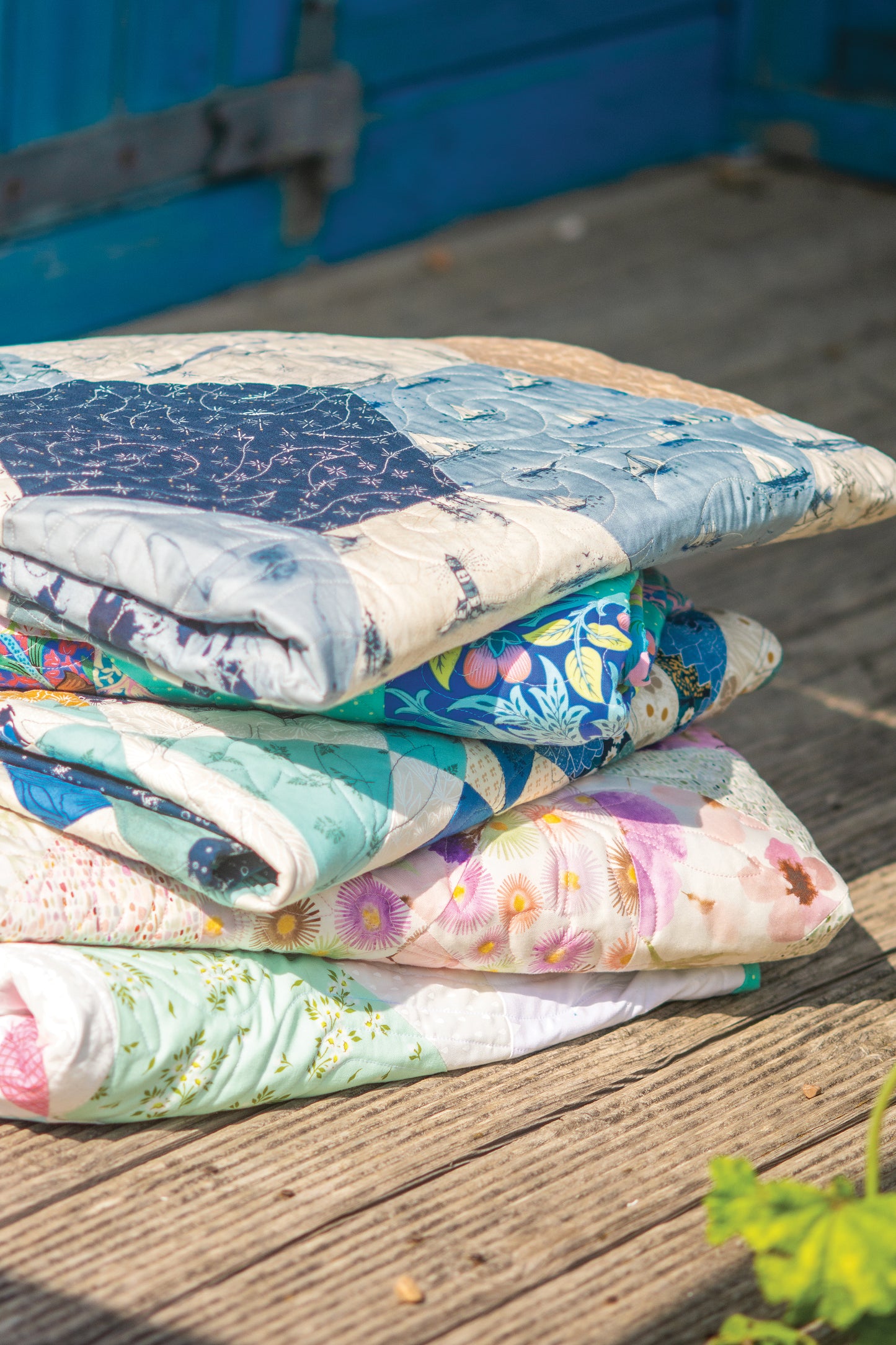 A vibrant stack of Layer Cake Quilts from Stash Books is neatly folded on a wooden deck under bright sunlight, with a blue door and soft green foliage blurred in the background.