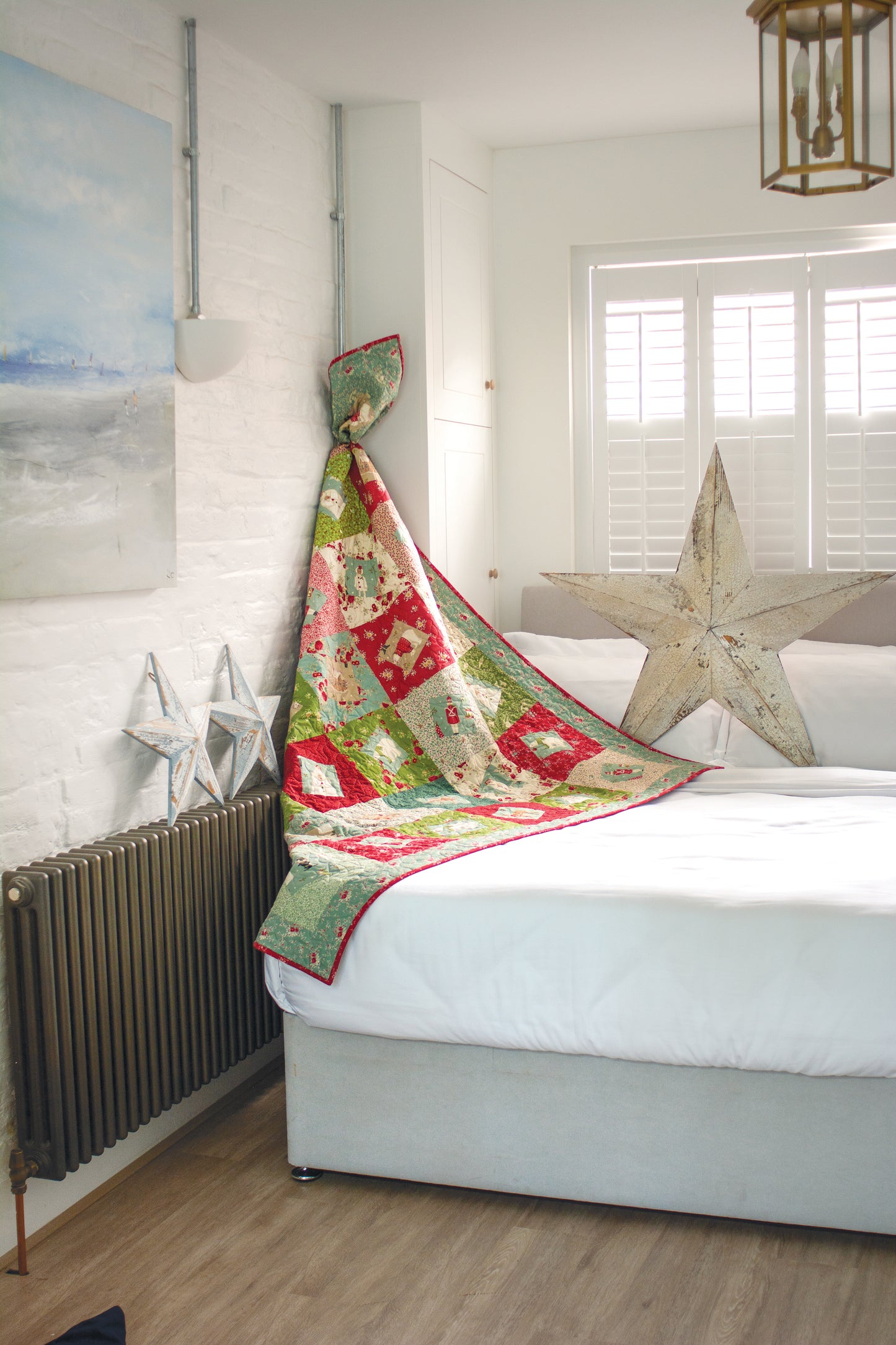 A Layer Cake Quilts creation from Stash Books, crafted with vibrant 10" precut squares, hangs off a neat bed in a bright room with white walls, wooden floor, metal star décor, and a large window with white shutters.