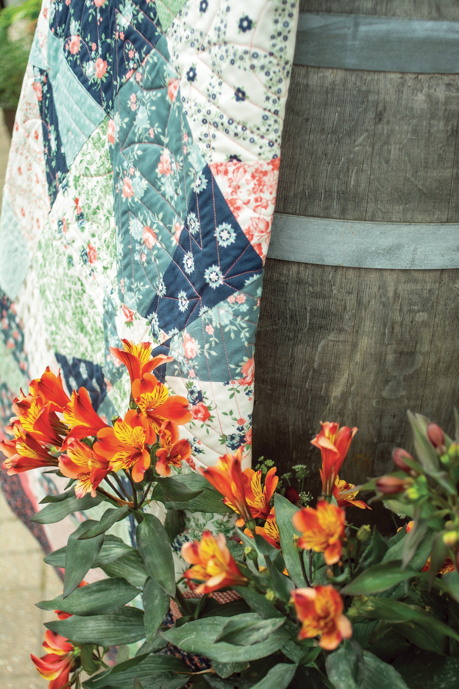 A Layer Cake Quilts design by Stash Books, crafted from 10" floral precut squares, drapes over a rustic wooden barrel beside bright orange and yellow flowers.