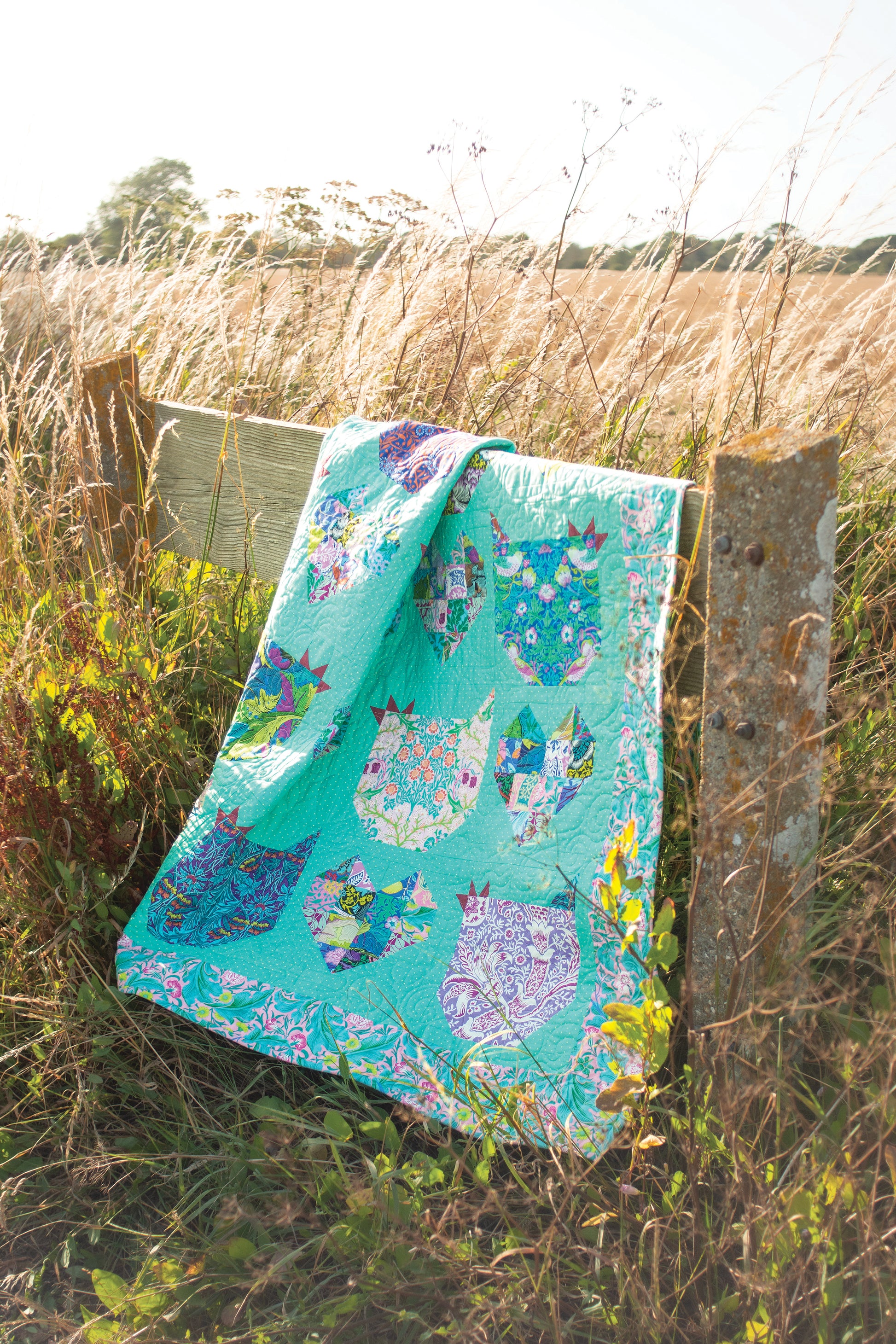 A vibrant quilt from Stash Books' Layer Cake Quilts, crafted with 10" precut squares and cat face designs, is draped over a wooden fence in a sunny grassy field.