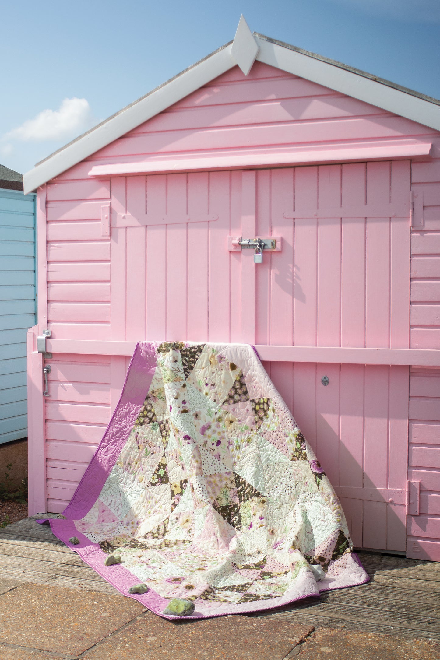 The Layer Cake Quilts from Stash Books, crafted from 10" precut squares in pastel purples, greens, and creams, is draped over a closed pink beach hut beneath a partly cloudy sky.