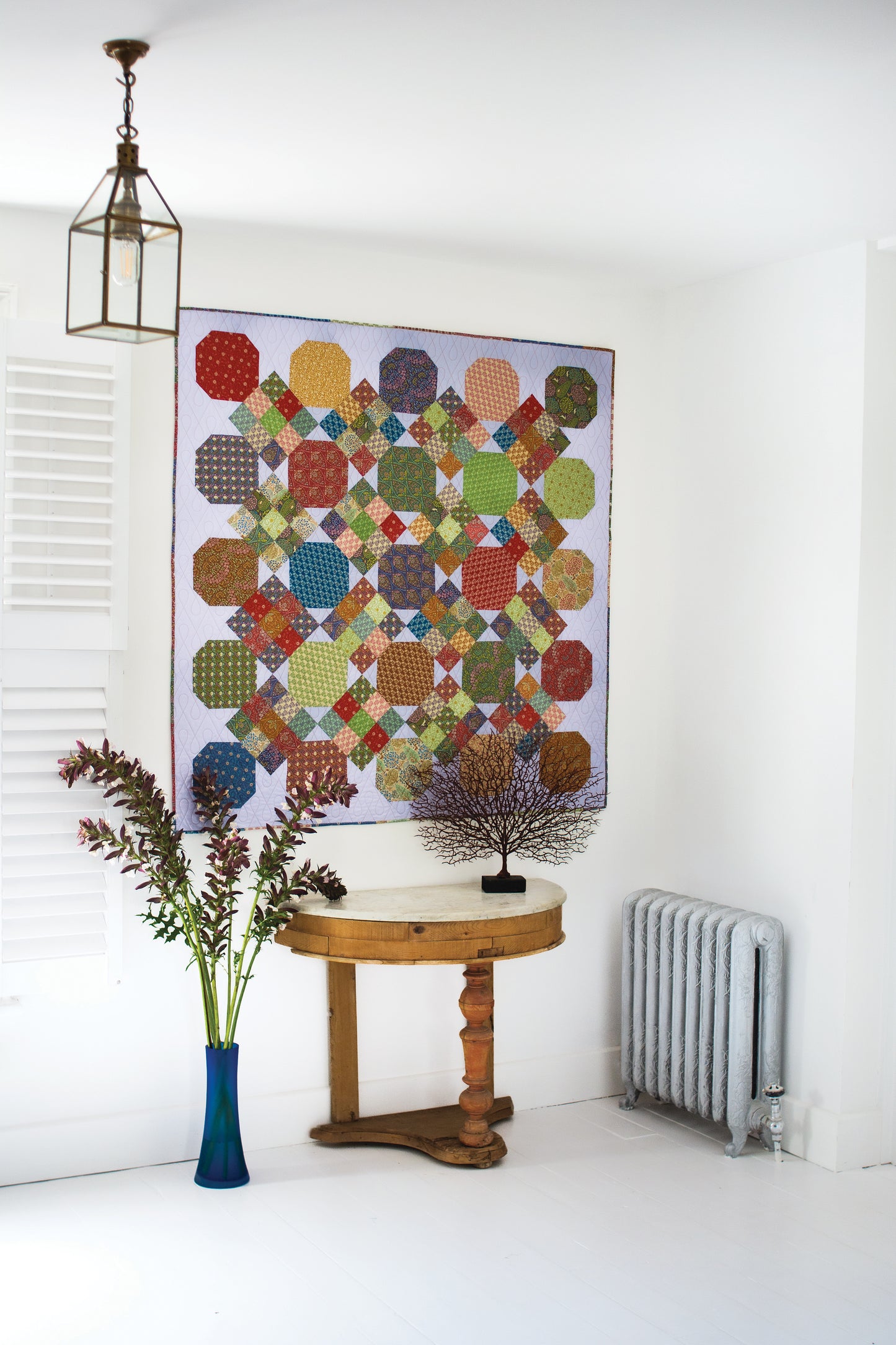 A vibrant Layer Cake Quilts by Stash Books hangs on a white wall above a small round table with a metal tree sculpture; beside it, a tall blue vase with leafy stems stands near a metal radiator.